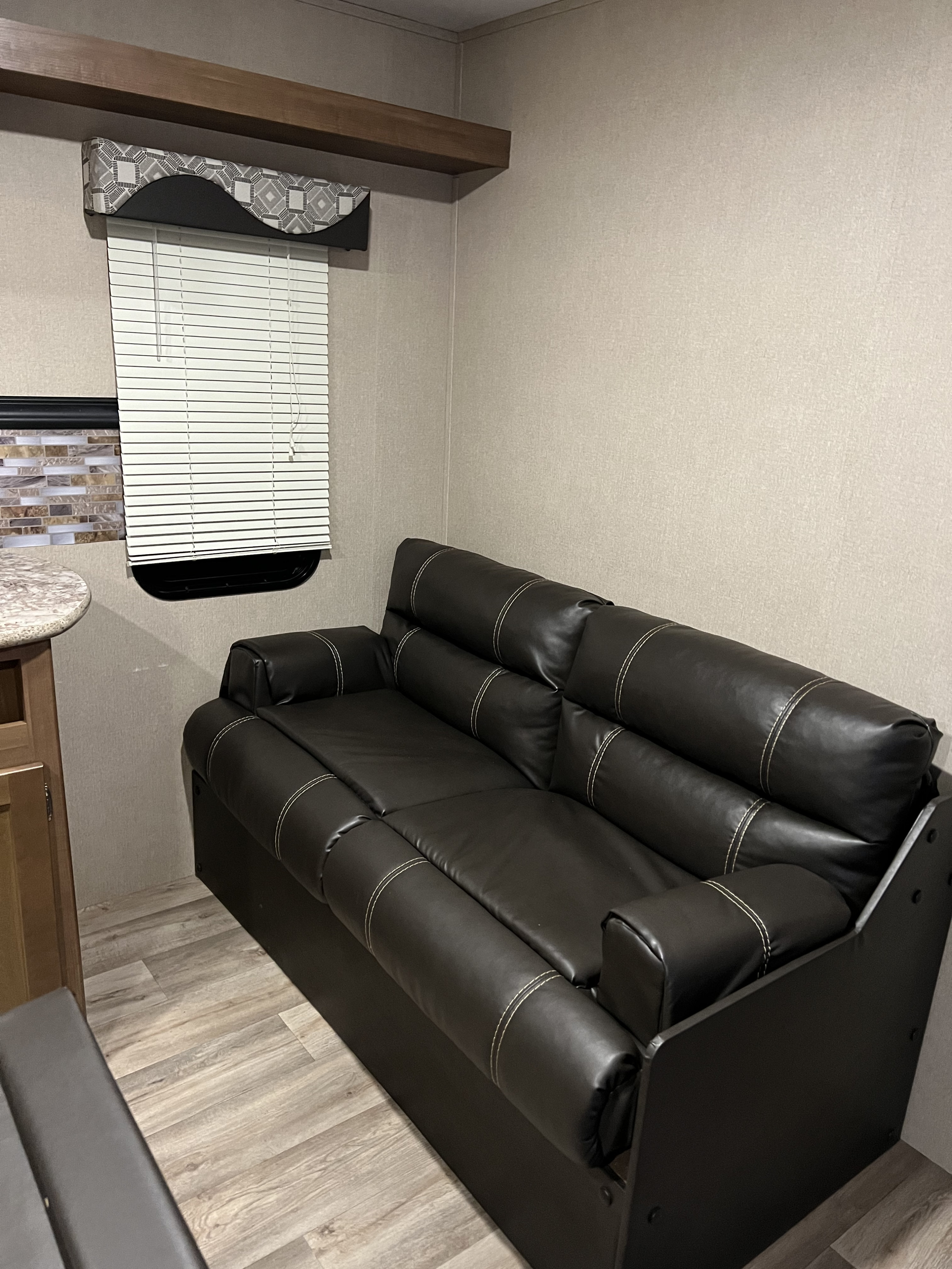 This cozy RV rental interior features a black sofa with light stitching, wood-look flooring, and a window with blinds.. Aspen Trail Aspen Trail Trailer 2018