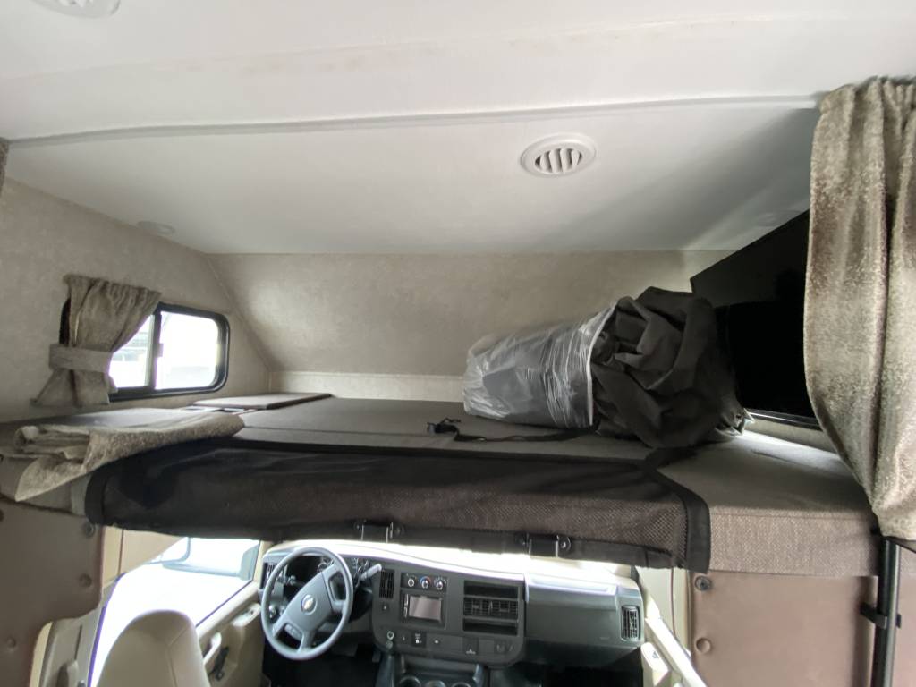 Inside an RV rental: a cozy cab-over bunk with bedding, driver's cockpit, window, and TV.. Forest River Sunseeker LE 2020