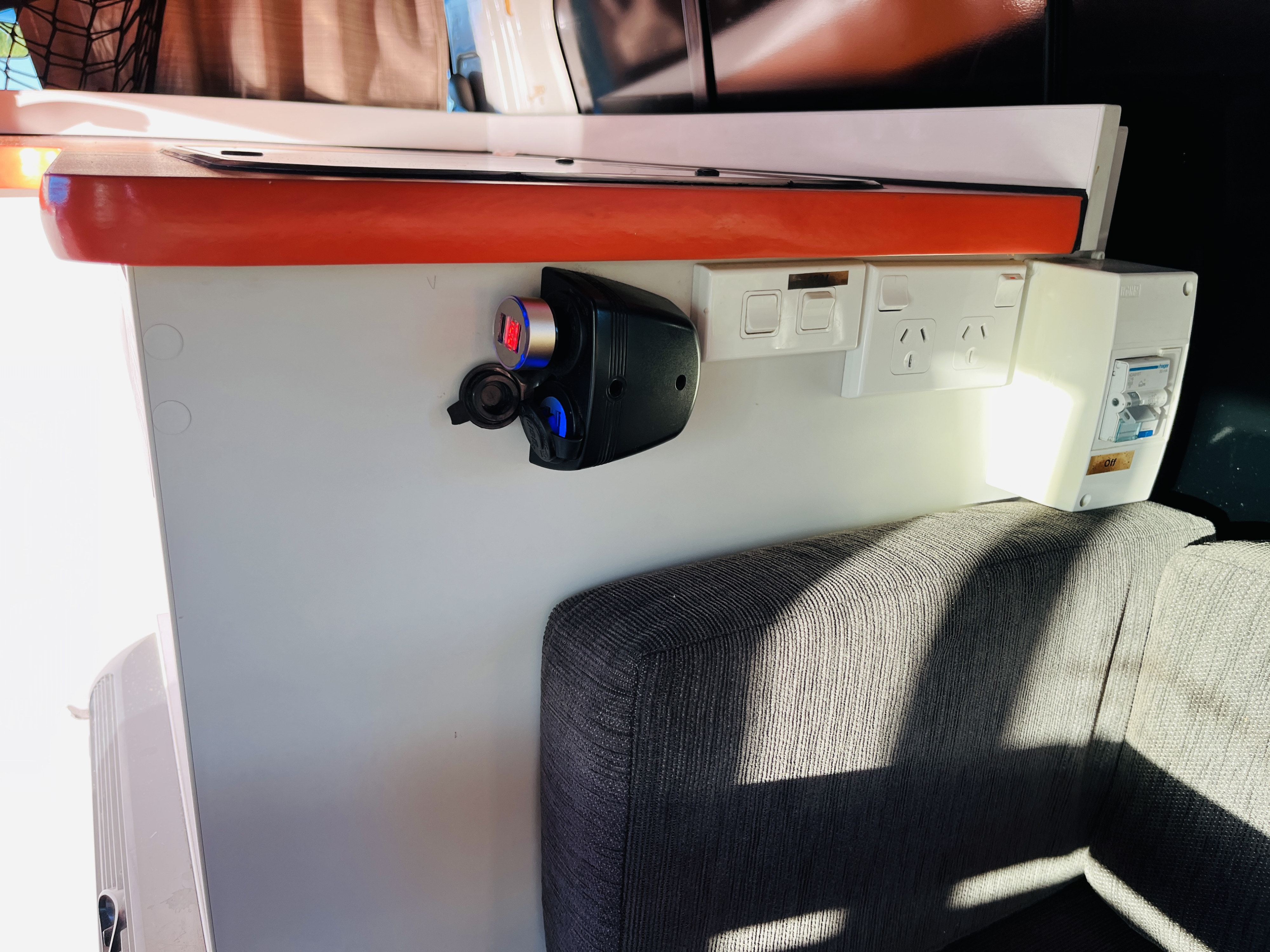 Australian power outlets, USB charger, and circuit breaker in an RV rental, mounted on a white panel above a grey bench seat.. Toyota Hiace 2009