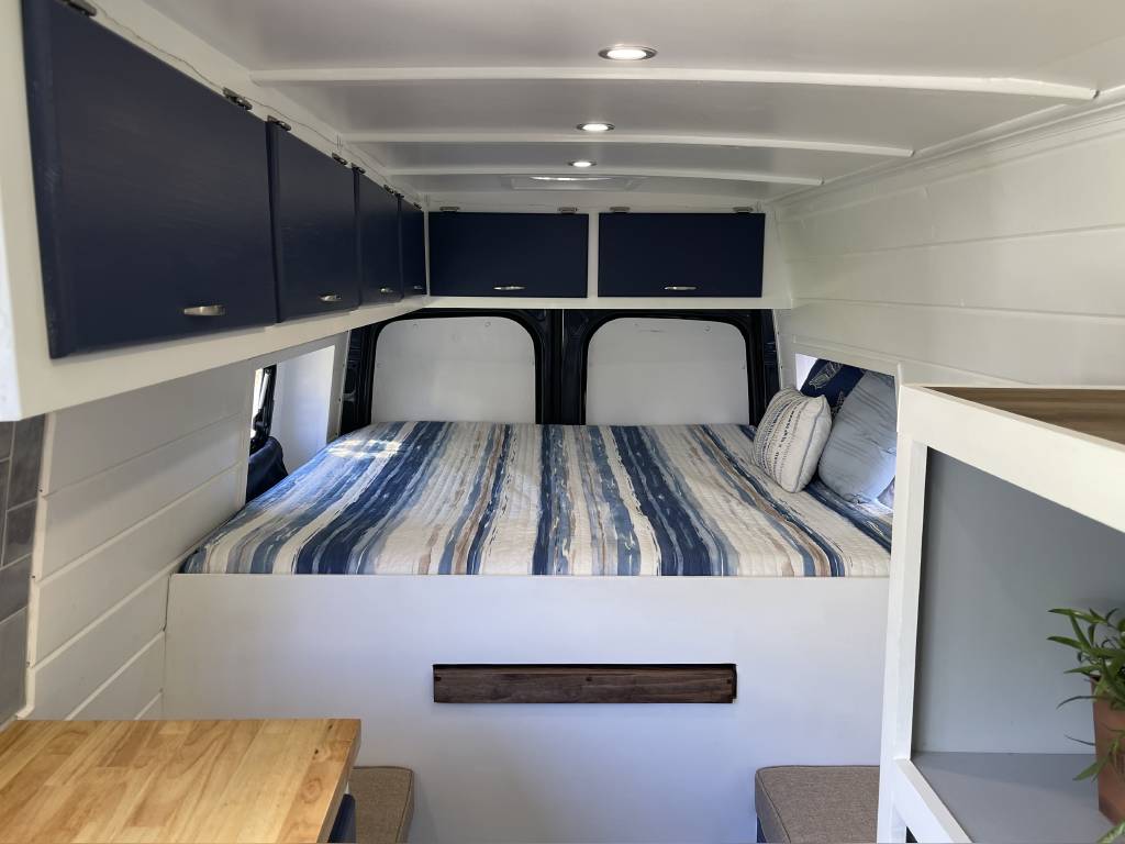 Inside a compact RV rental: a comfy bed with striped bedding, blue cabinets, and white paneling. Ideal for road trips.. Mercedes Benz Sprinter 2500 2020