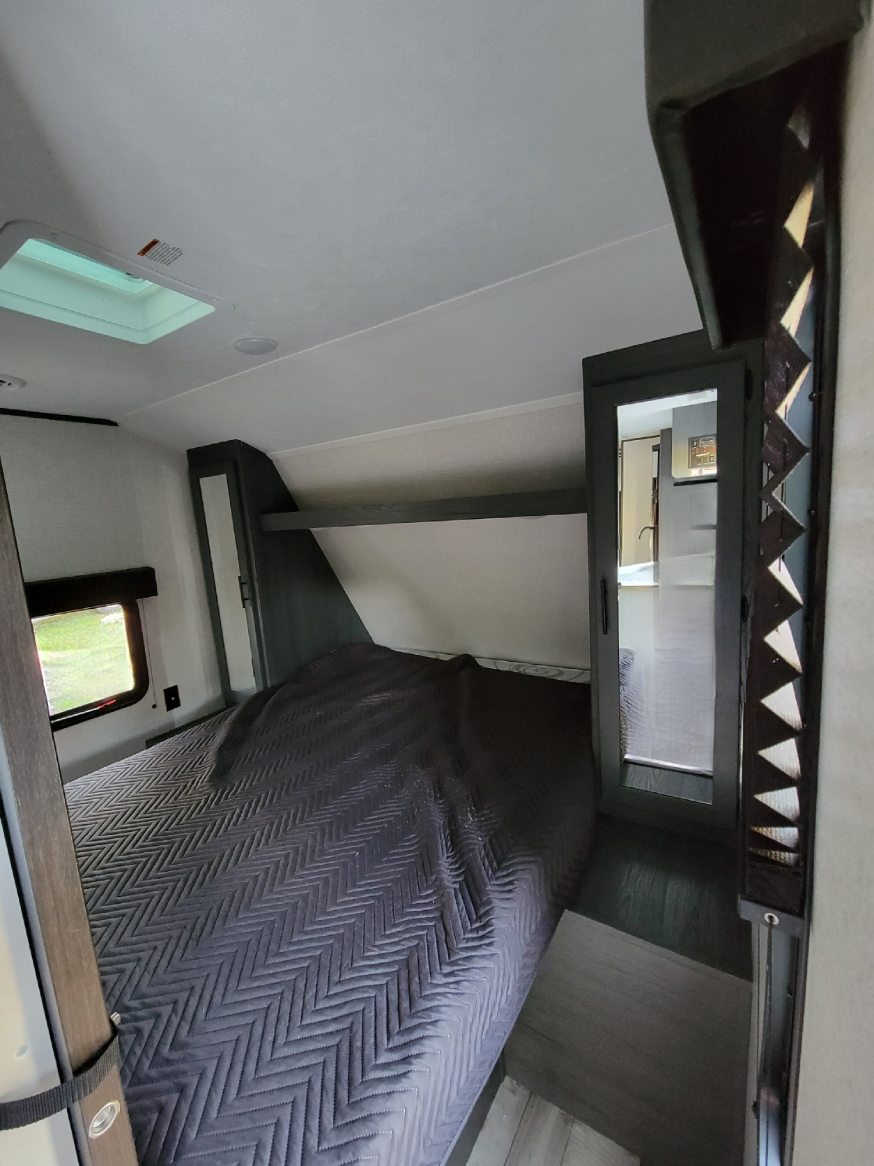 Modern RV rental bedroom with a gray quilted bed, overhead bunk, window, and cabinet reflecting another room.. Dutchman Aspen Trail 2022