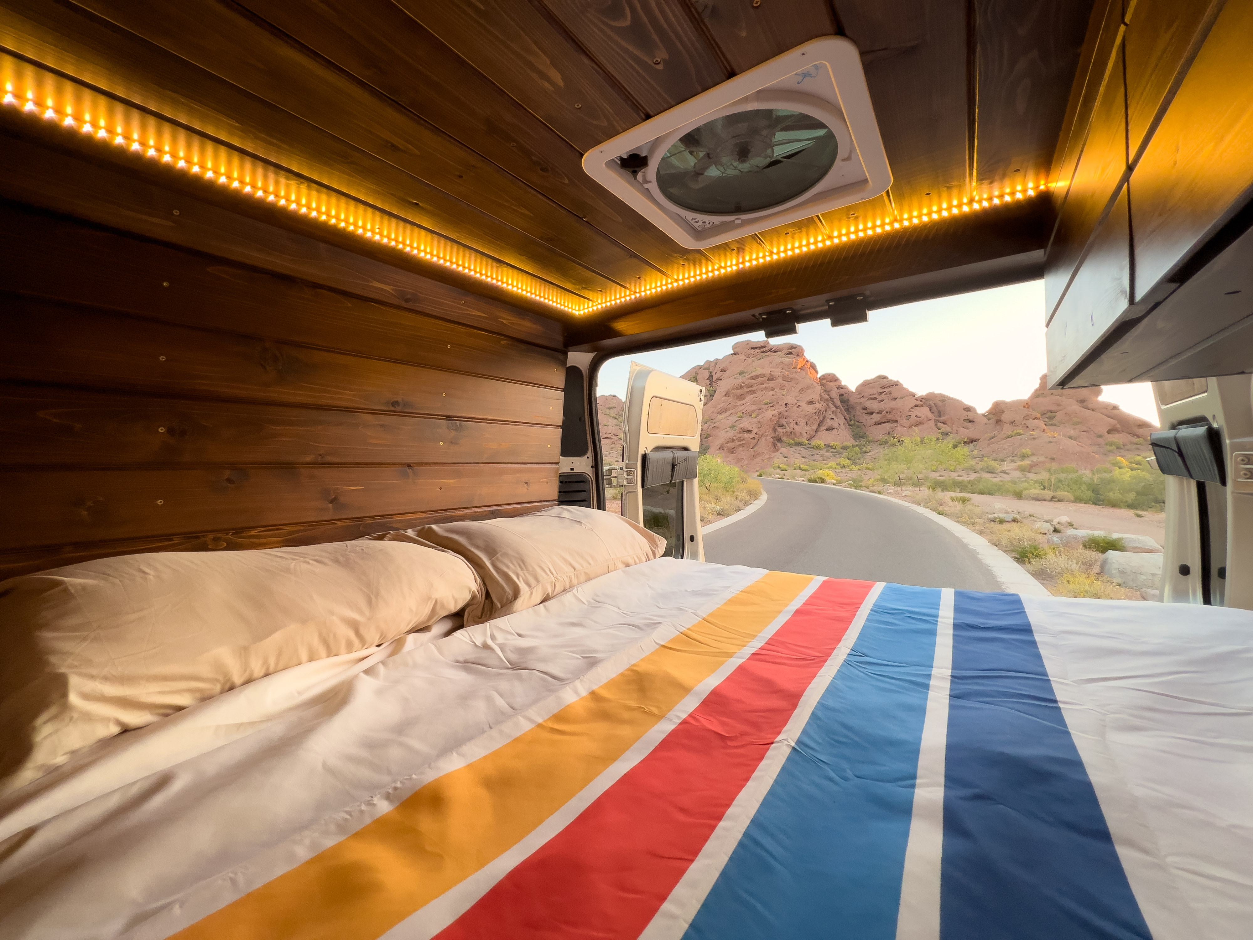 Cozy RV rental bedroom with colorful bedding & warm LED lights, opening to a scenic desert road and red rock mountains.. RAM Promaster 2021
