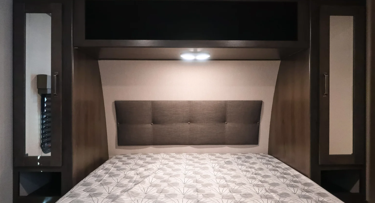 Relaxing RV rental bedroom featuring a padded headboard, overhead storage, and compact side cabinets.. Grand Design Xplor 2022