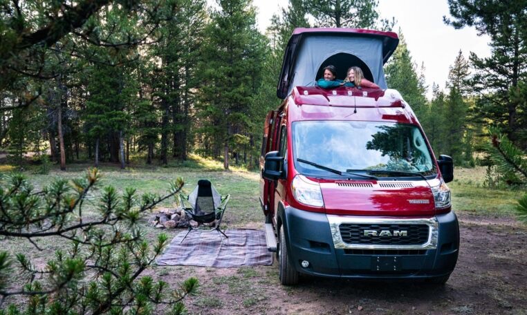 Two people enjoy an RV rental with a pop-top roof at a scenic forest campsite, complete with a fire pit and chair.. Winnebago Solis 2021
