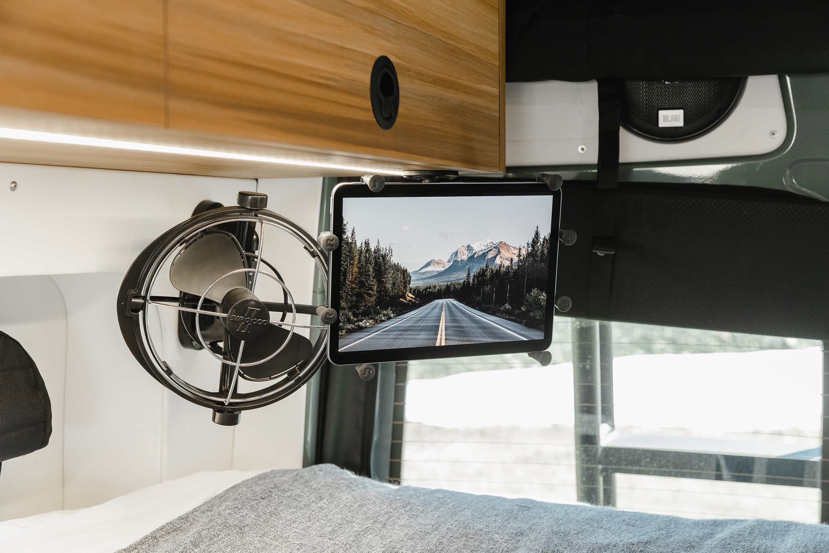 RV rental interior: overhead cabinet with light, fan, tablet showing scenic road. Cozy bed, window in background.. Mercedes-Benz Sprinter 2021