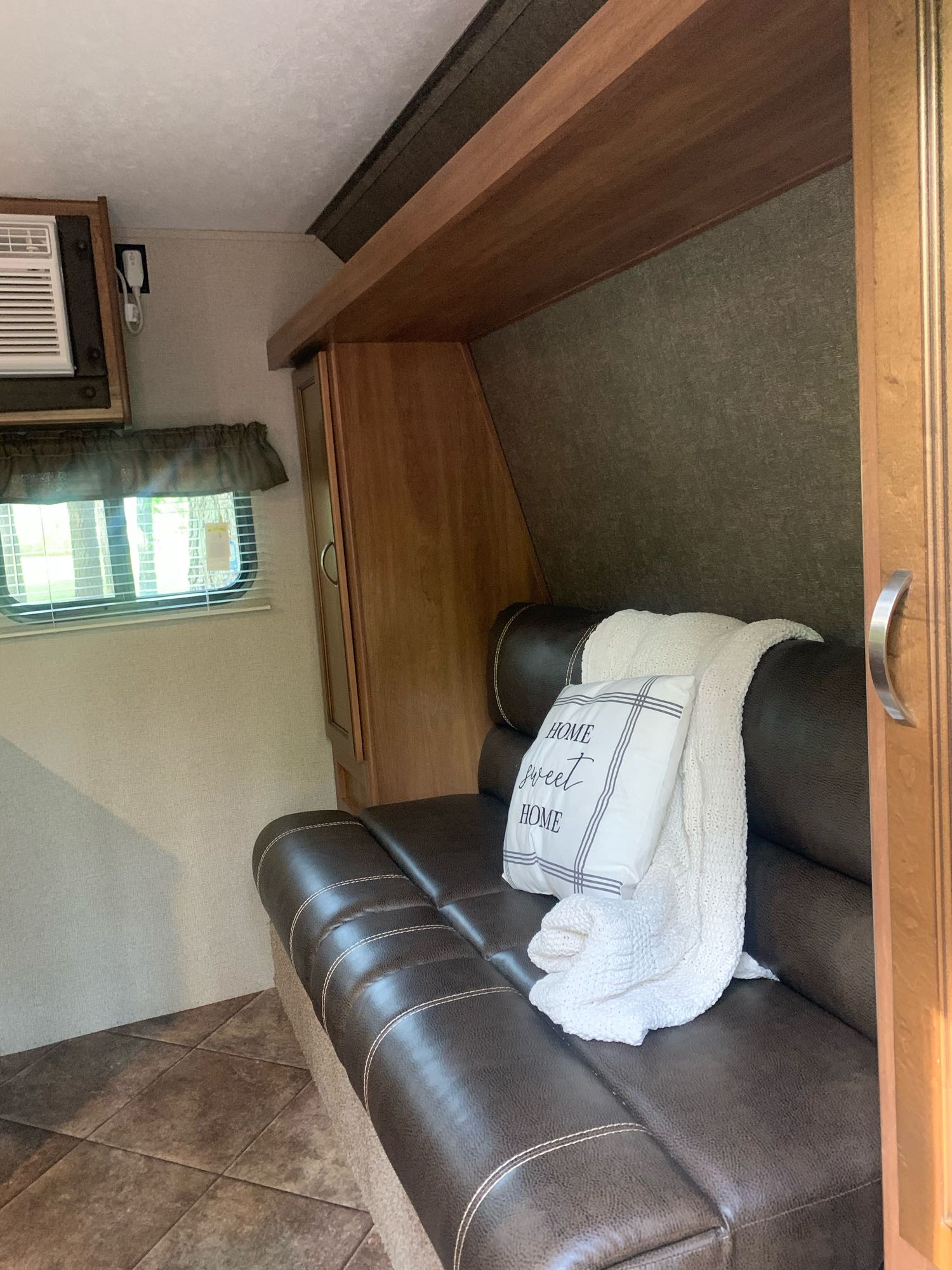 Cozy RV rental interior featuring a brown sofa, "Home Sweet Home" pillow, window with blinds, AC, and wood cabinetry.. Keystone RV Summerland Mini 2018