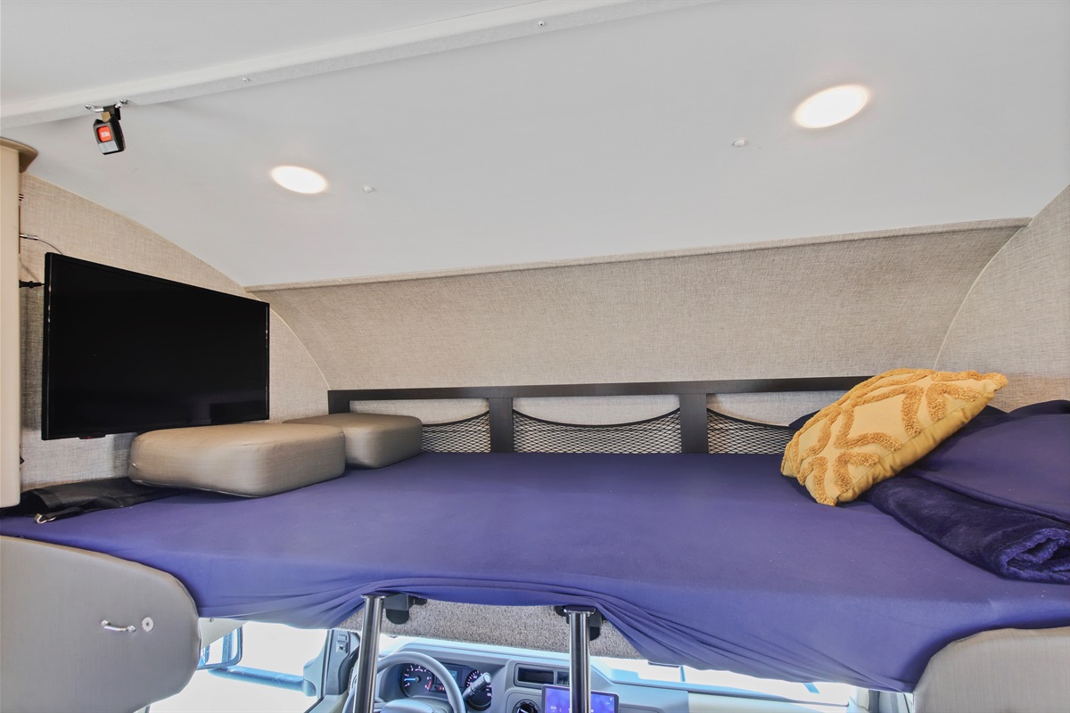 RV rental's spacious cab-over bunk with a mounted TV, purple bedding, and headboard storage nets. A comfy RV sleeping area.. Jayco Redhawk 2022