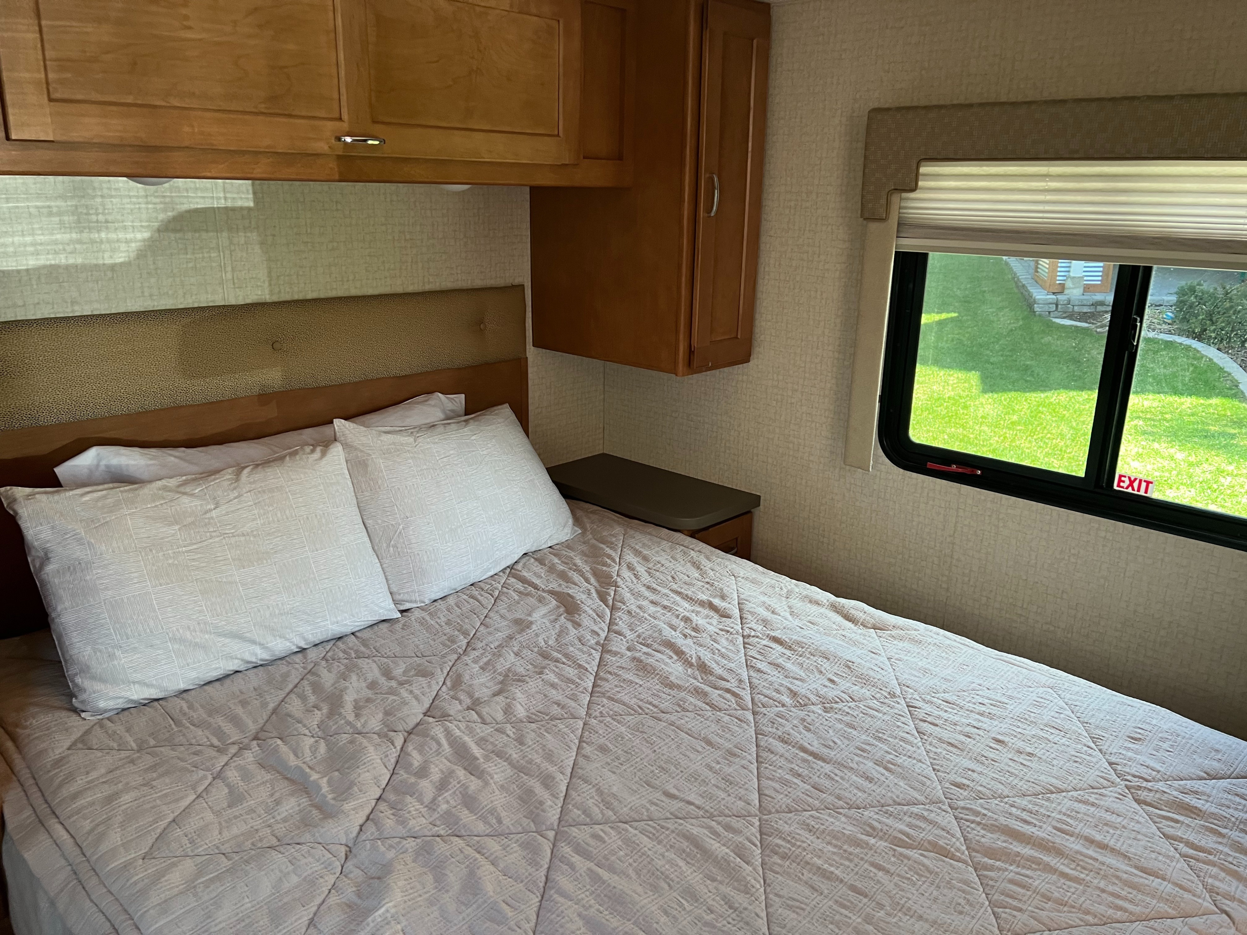 Cozy rv rental bedroom featuring a comfortable bed with light bedding, overhead cabinets, and a window revealing a green landscape outside.. Winnebago Minnie Winnie 2017