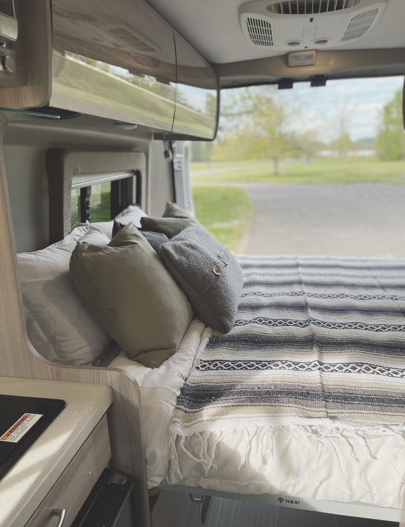 Cozy RV rental interior with a bed, neutral pillows, and a striped blanket facing a scenic nature view. (THOR RV visible).. Thor Sequence 2021