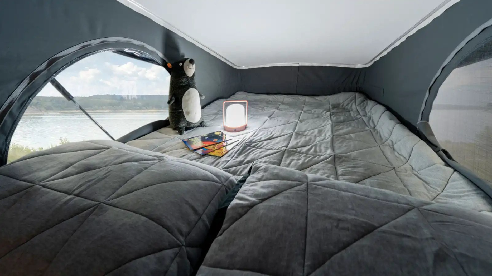 Cozy rv rental pop-top bed with grey quilted bedding, a lamp, and books, offering stunning lake views from the window.. Winnebago Solis 2022