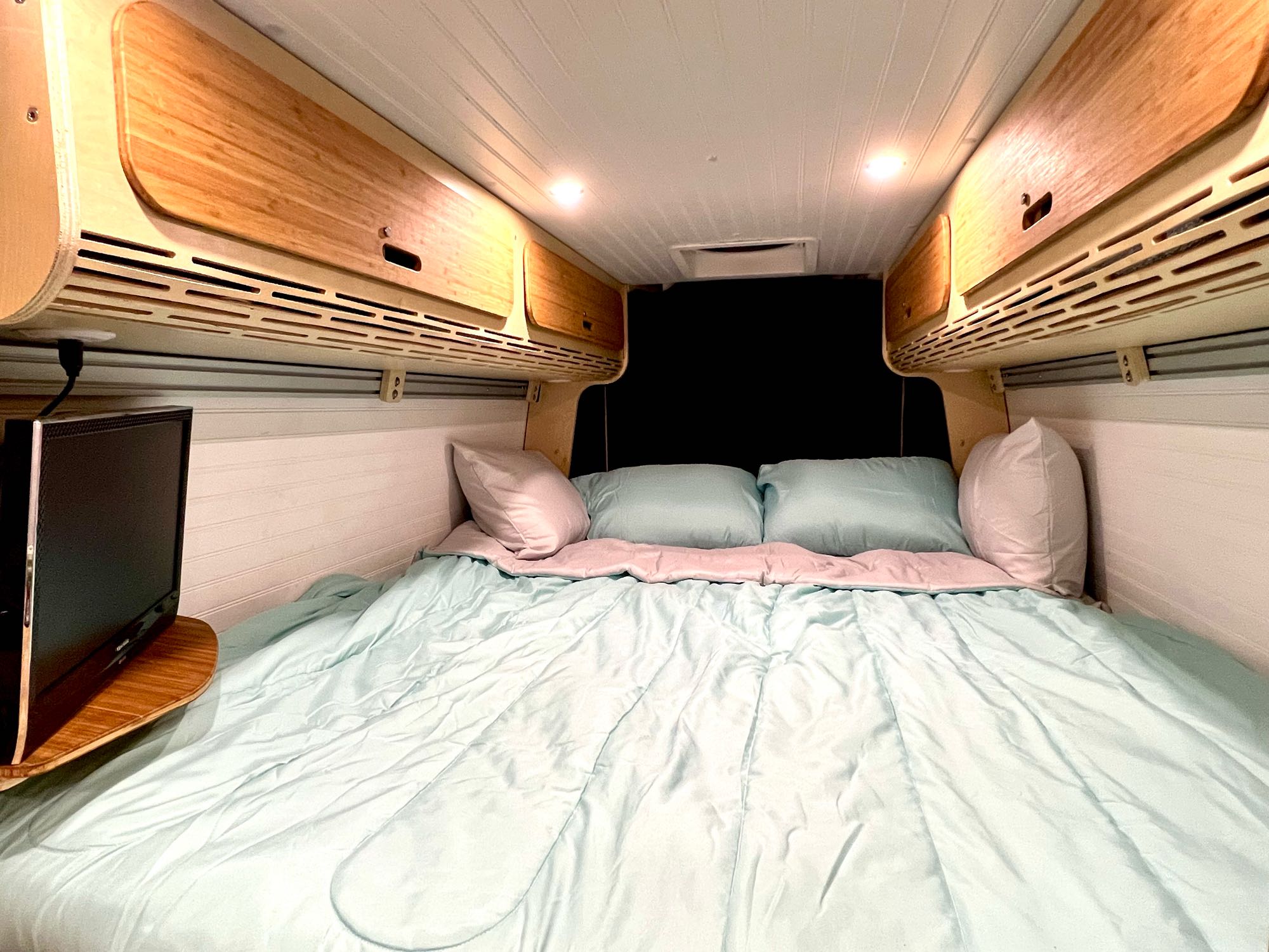 Cozy RV rental interior: A made bed with light blue comforter, overhead wood cabinets, TV, and white paneling.. Mercedes-benz Sprinter 2016