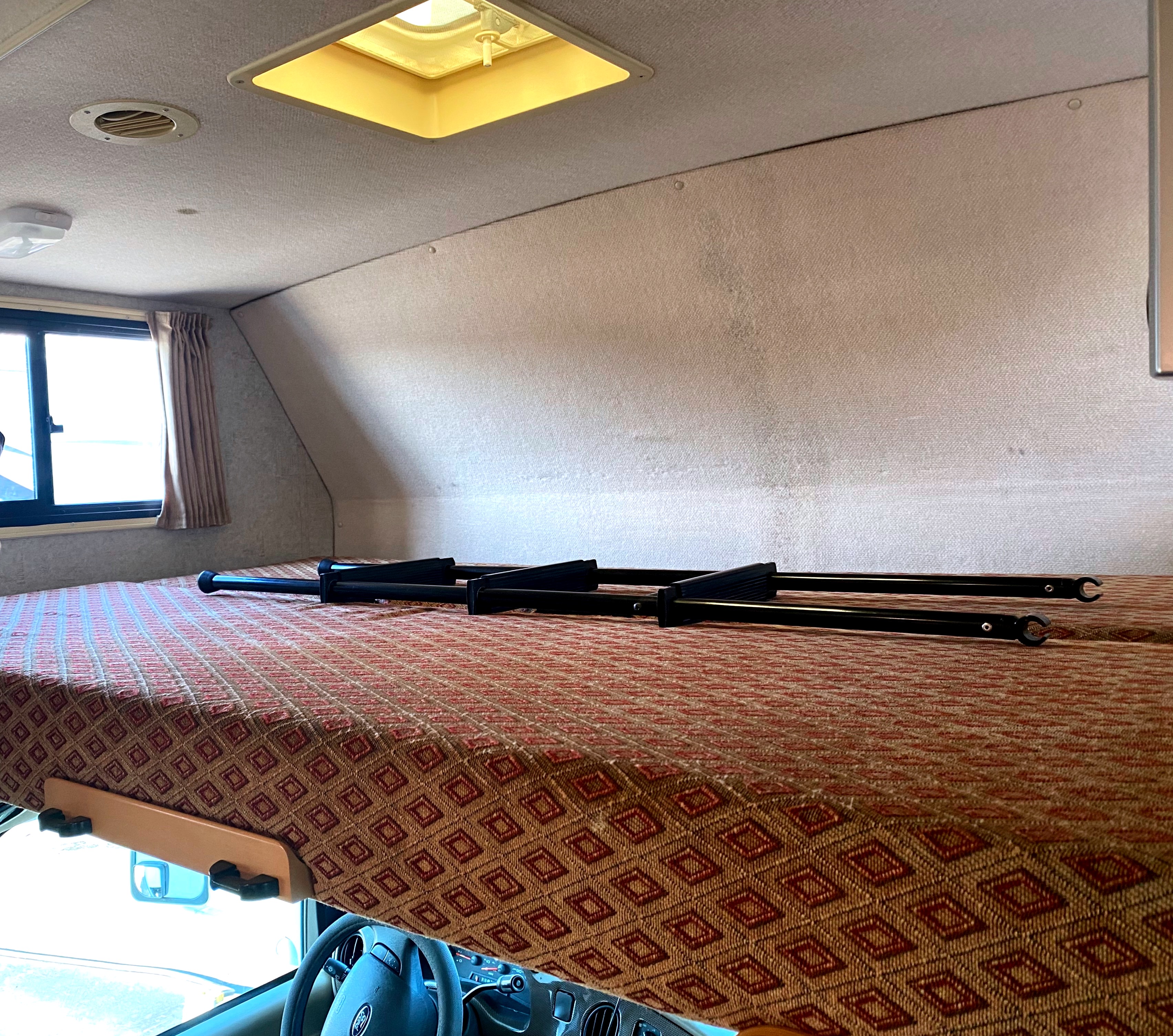 Inside an RV rental: a large cab-over bunk with a patterned mattress and disassembled ladder, overlooking the driver's cabin.. Winnebago Outlook 2007