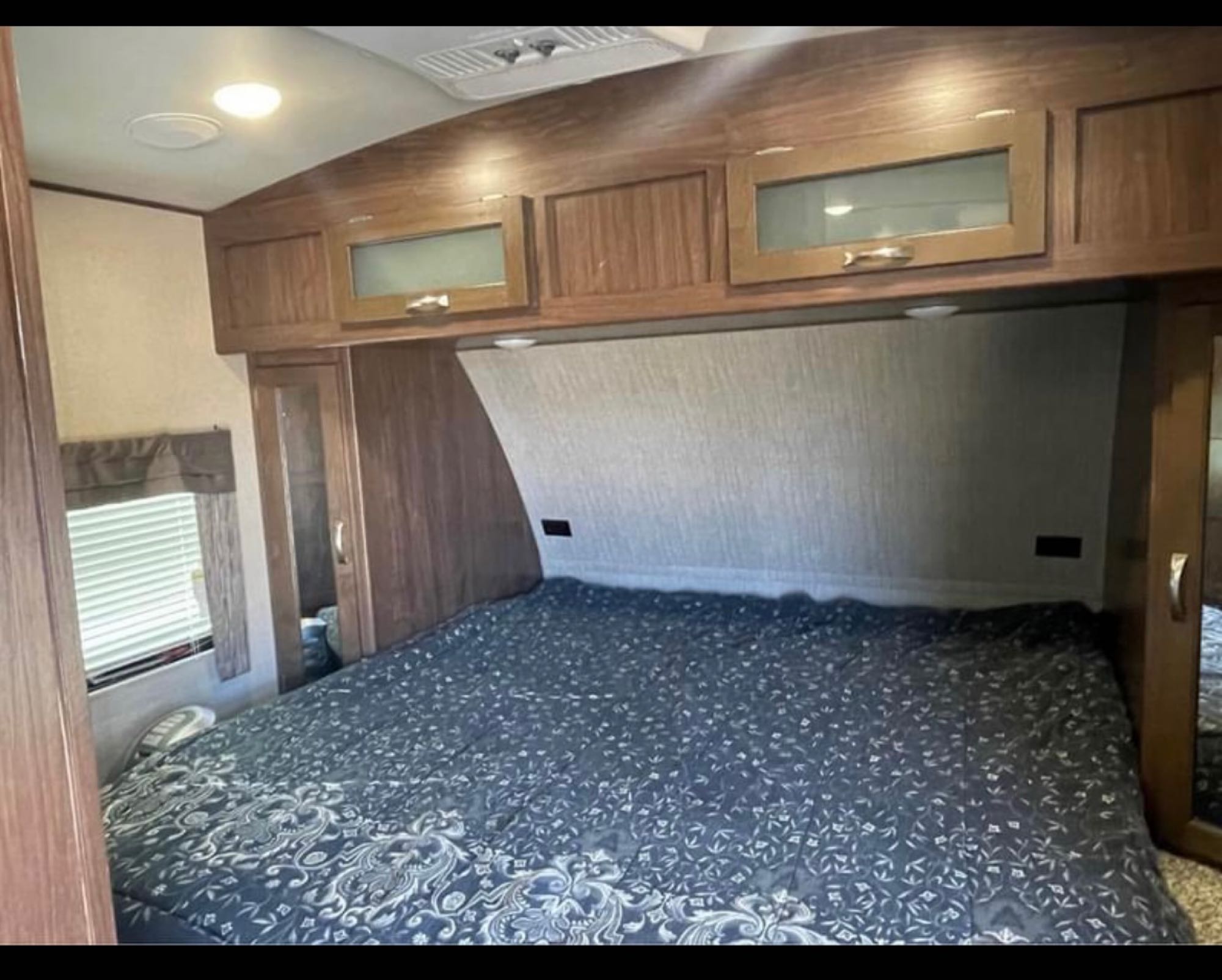 This RV rental features a cozy bedroom with a large bed, wooden overhead cabinets, side wardrobes, and a window with blinds.. Dutchman Aerolite Luxury Class 2017