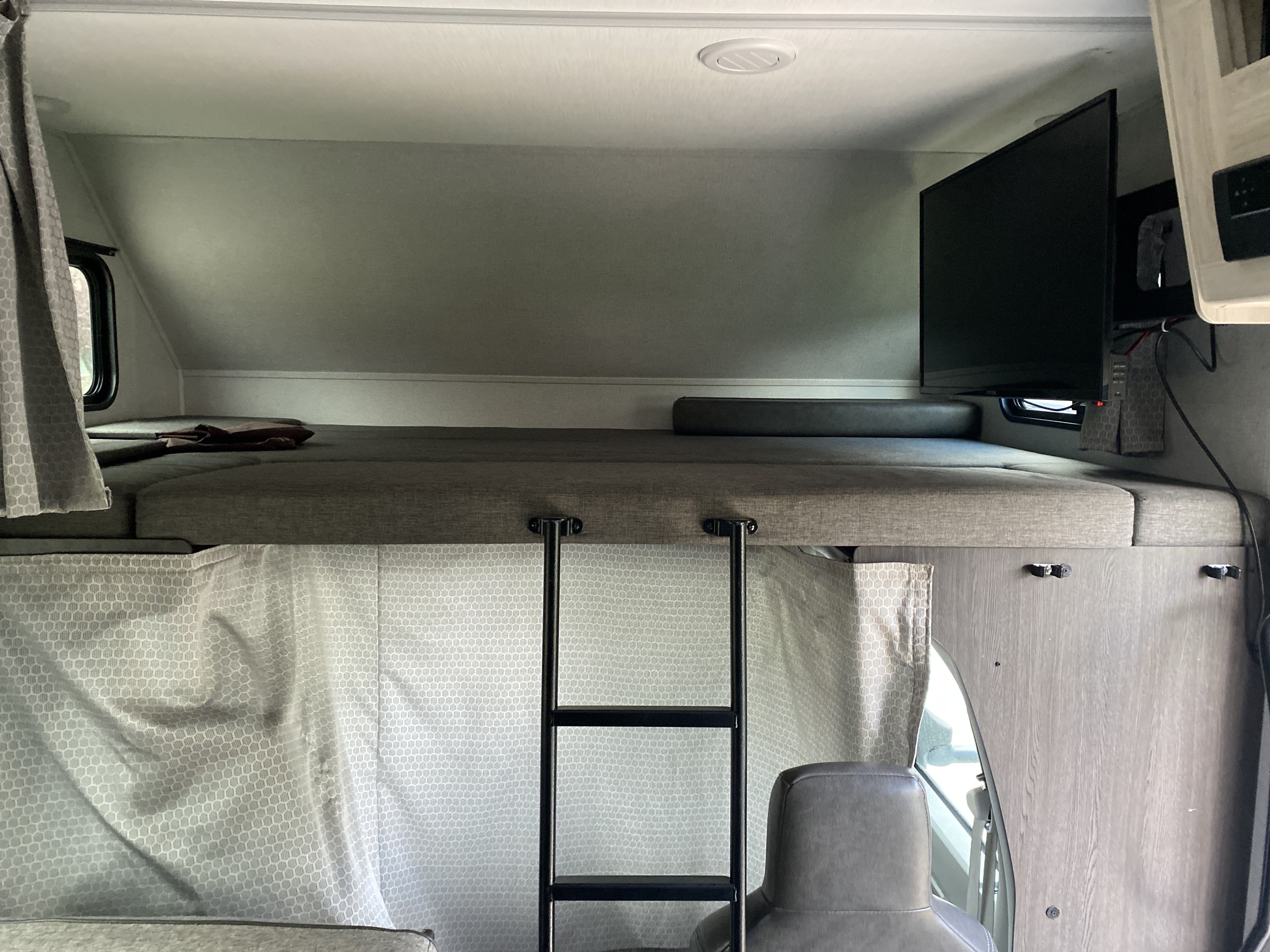 An RV rental over-cab bunk features a bed, TV, ladder, and patterned window curtains.. Forest River Sunseeker LE 2021