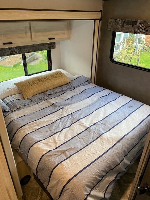 Inside an RV rental: a compact bedroom with a bed, striped bedding, overhead cabinets, and windows overlooking nature.. Forest River 2550DSLE 2022