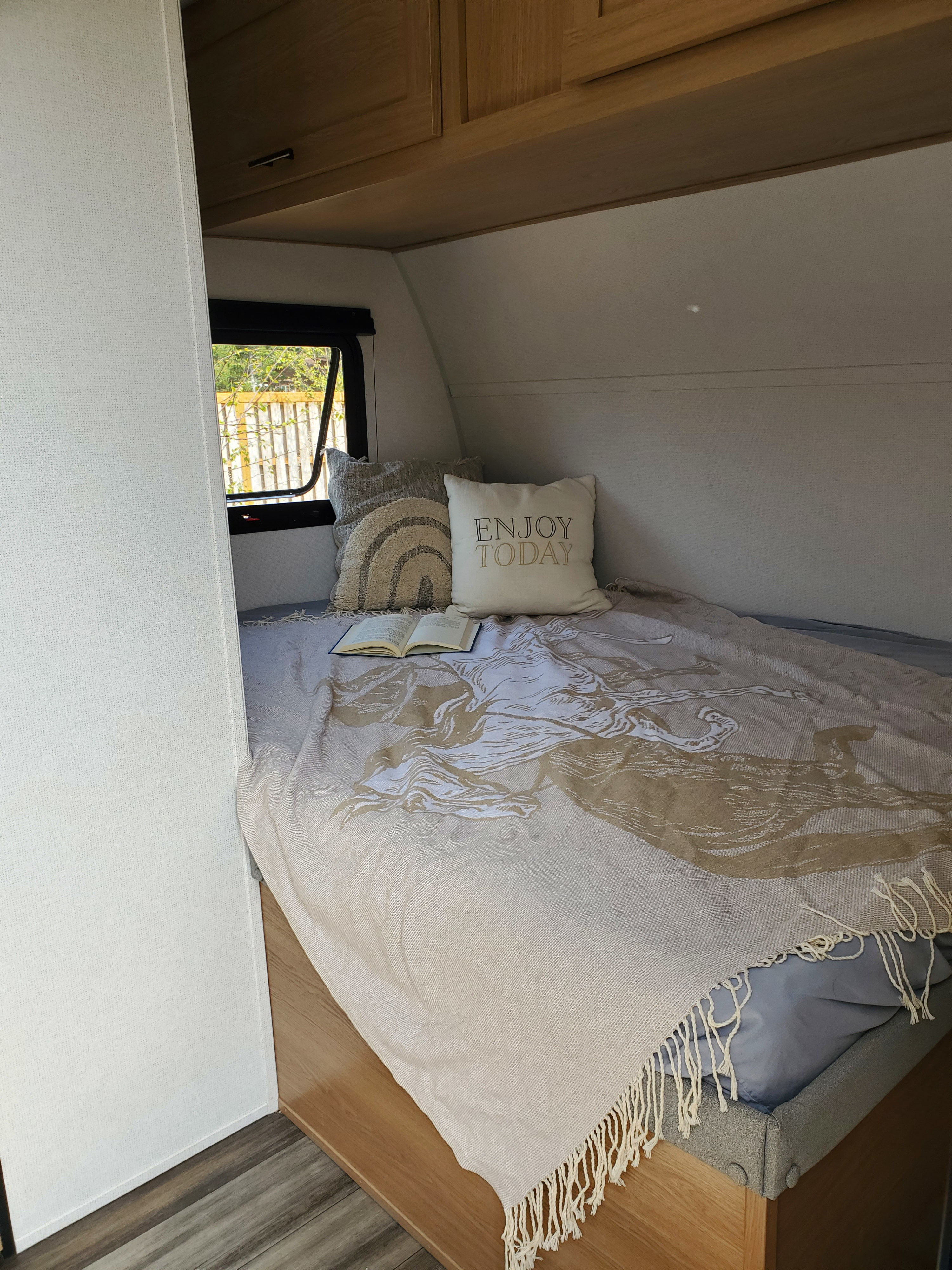Cozy RV rental bedroom with a comfortable bed, 'Enjoy Today' pillow, open book, and a scenic window view.. Forest River Avenger LT 16RD 2022