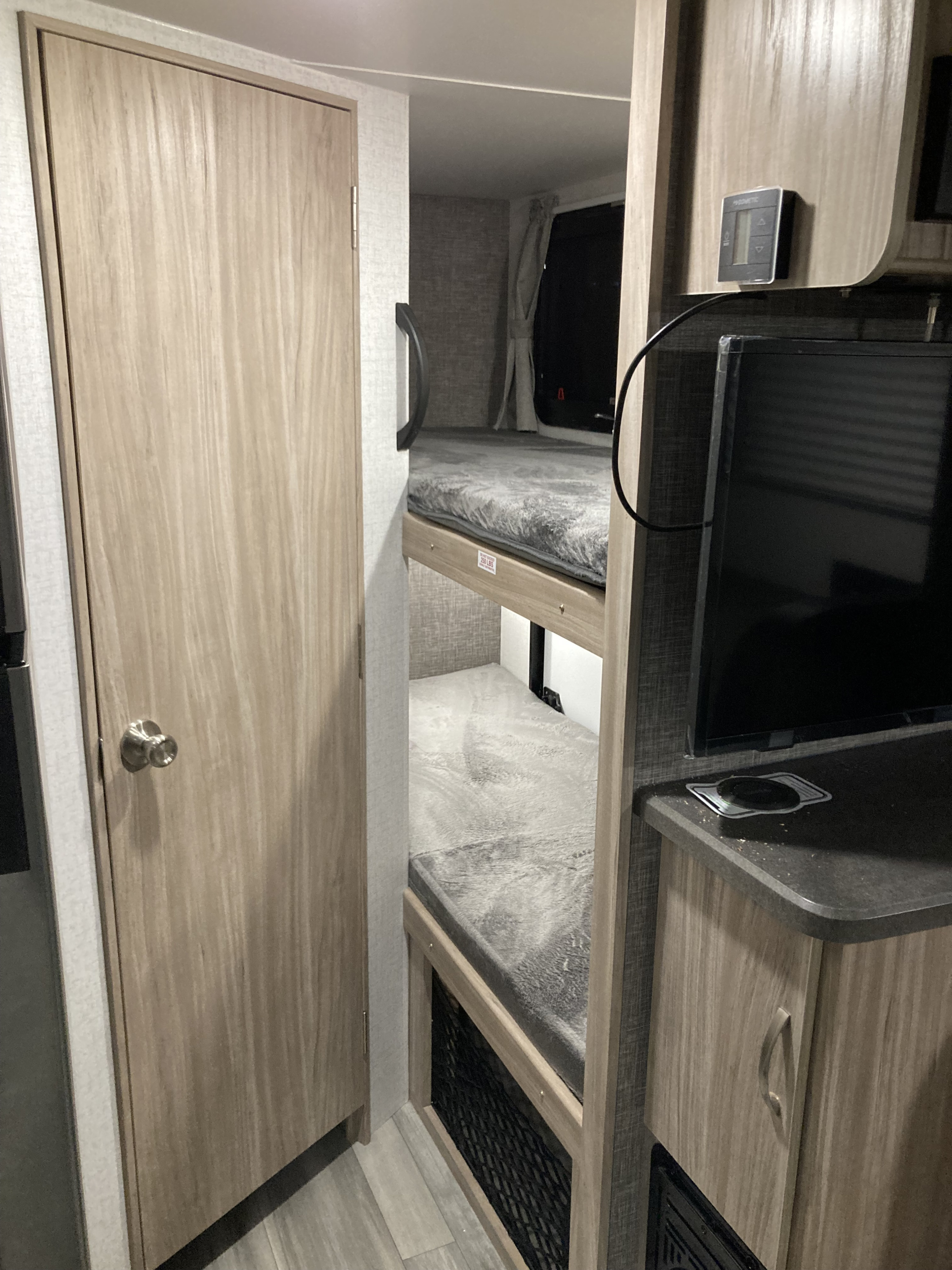 Experience a comfortable **rv rental**: light wood cabinetry, a door, and cozy bunk beds with soft grey blankets and TV area.. Winnebago Micro Minnie 2022