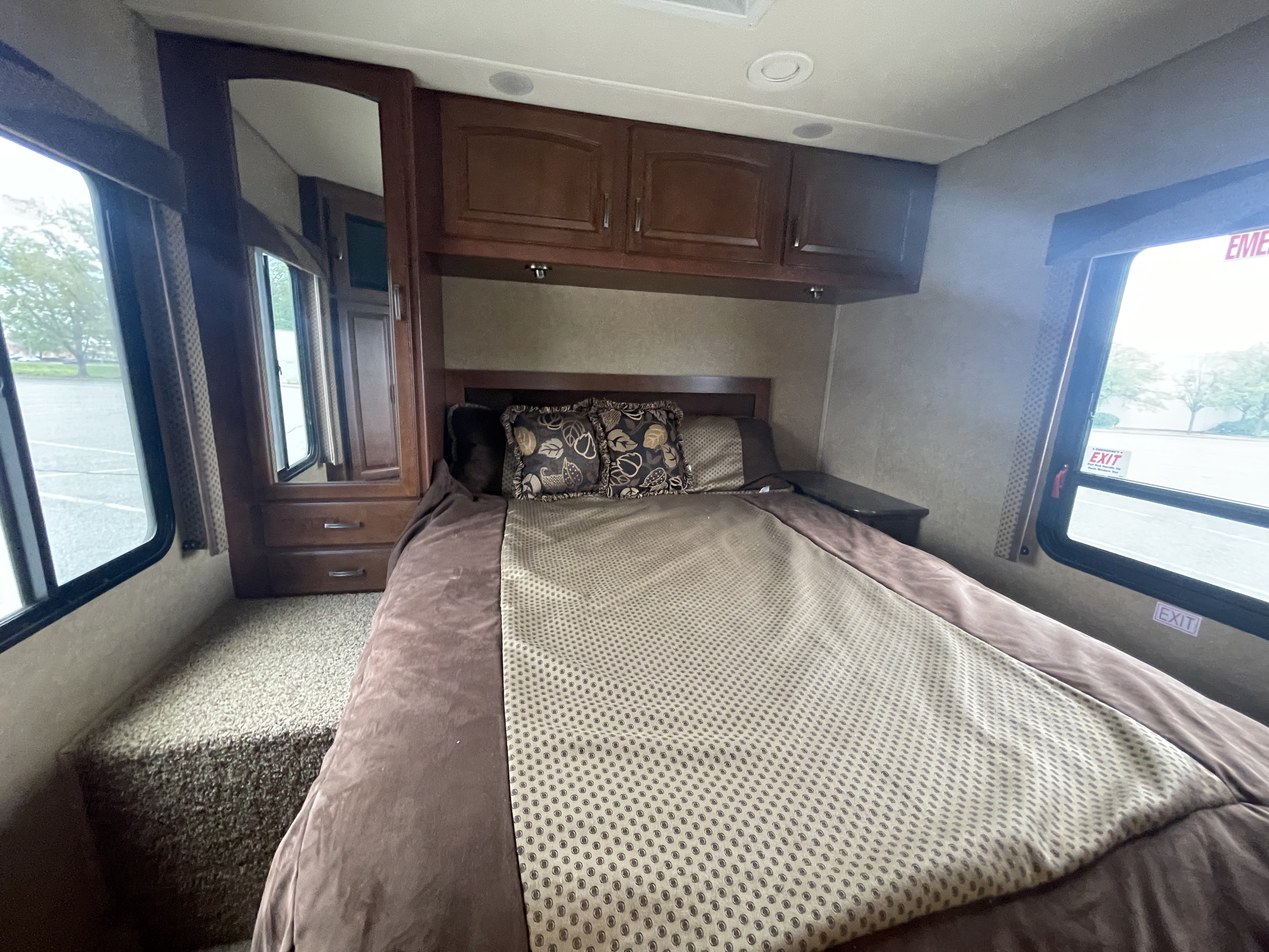 Cozy RV rental bedroom featuring a made bed, overhead cabinets, and large windows revealing a parking lot outside.. Jayco Greyhawk 2014