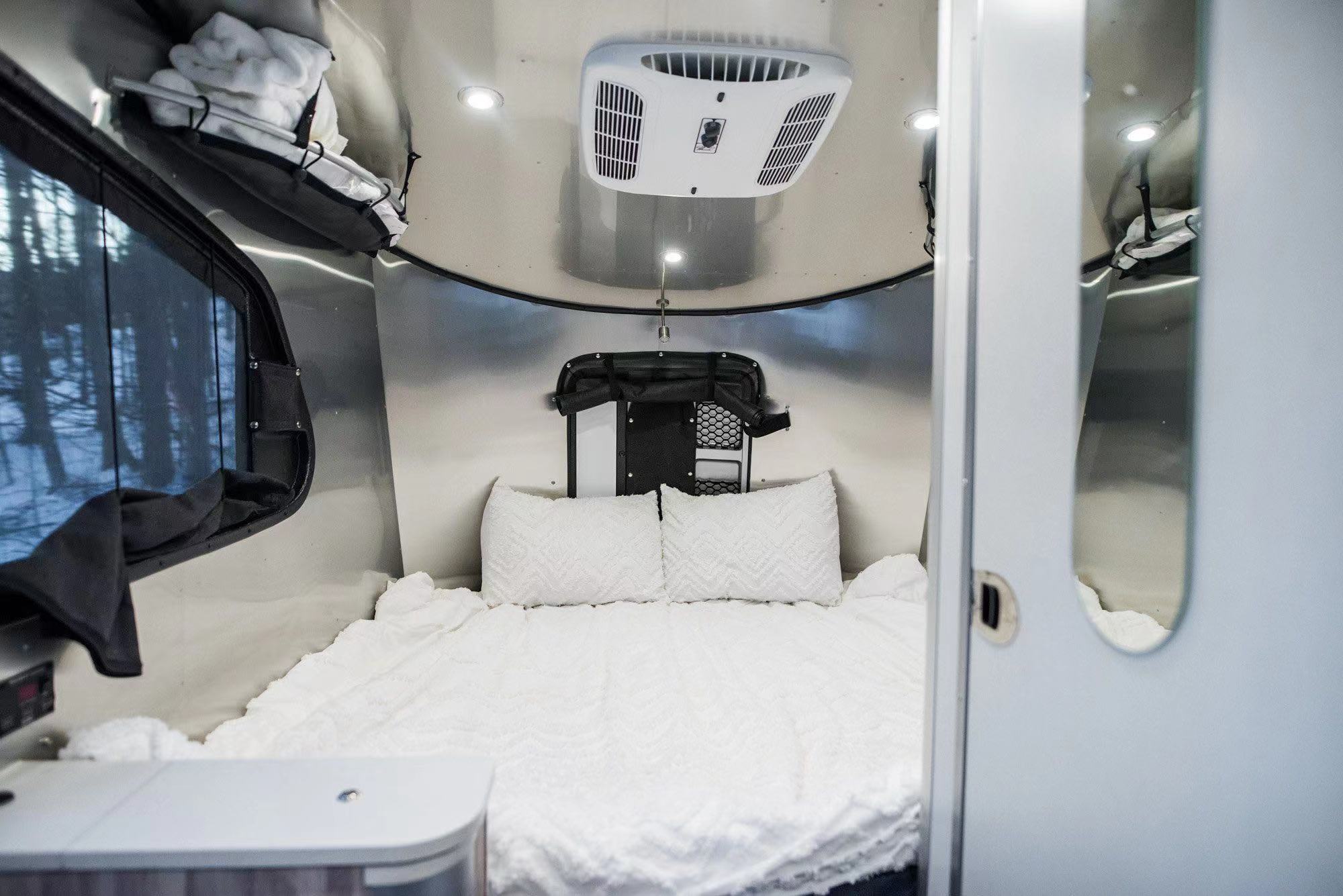 Inside this cozy RV rental: a plush bed with white linen, AC unit, & window view of a snowy winter forest.. Airstream Basecamp 2017