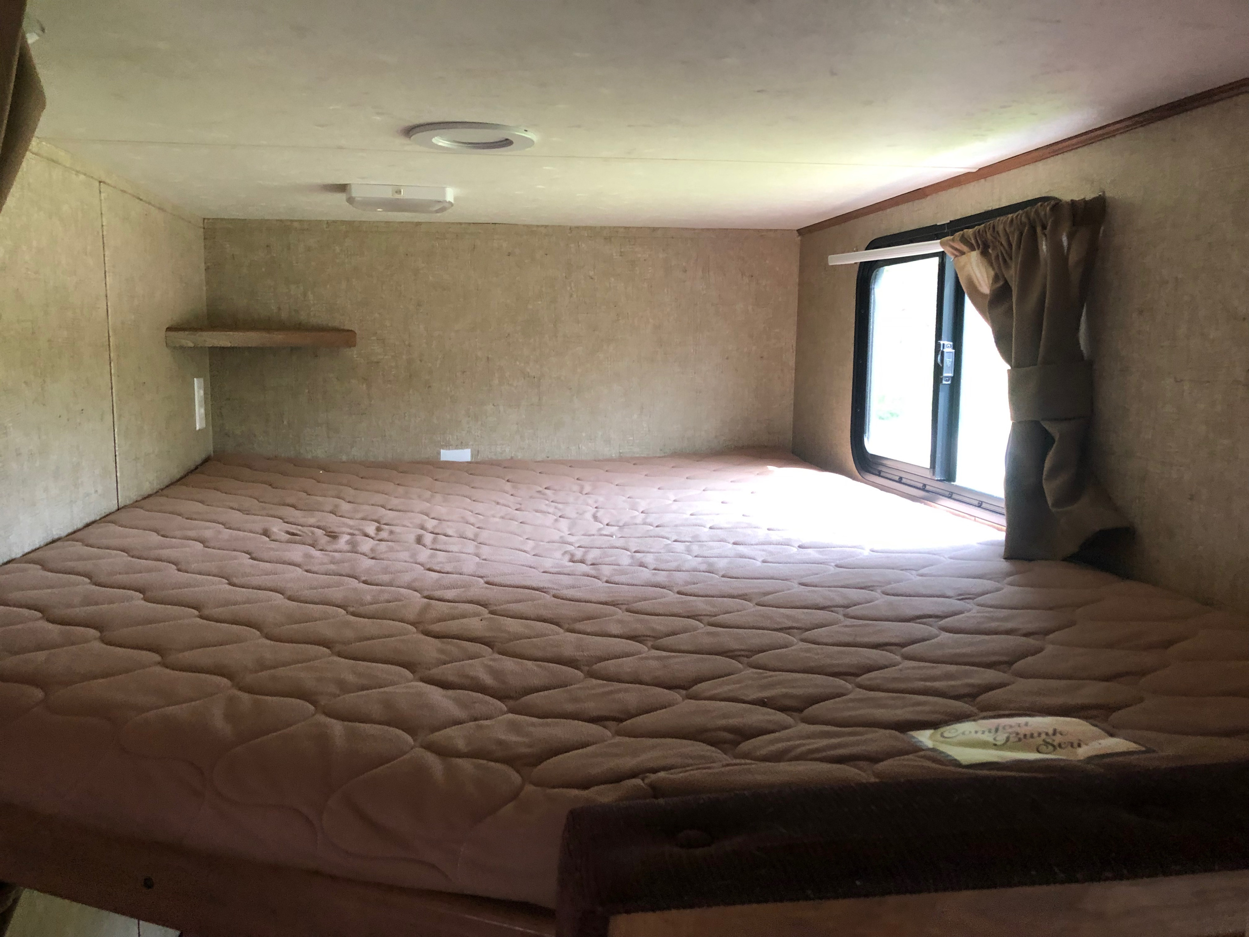 Cozy RV rental sleeping area featuring a wide quilted mattress, window with curtain, and wall shelf. Perfect for camping trips.. Coachmen Apex 2016