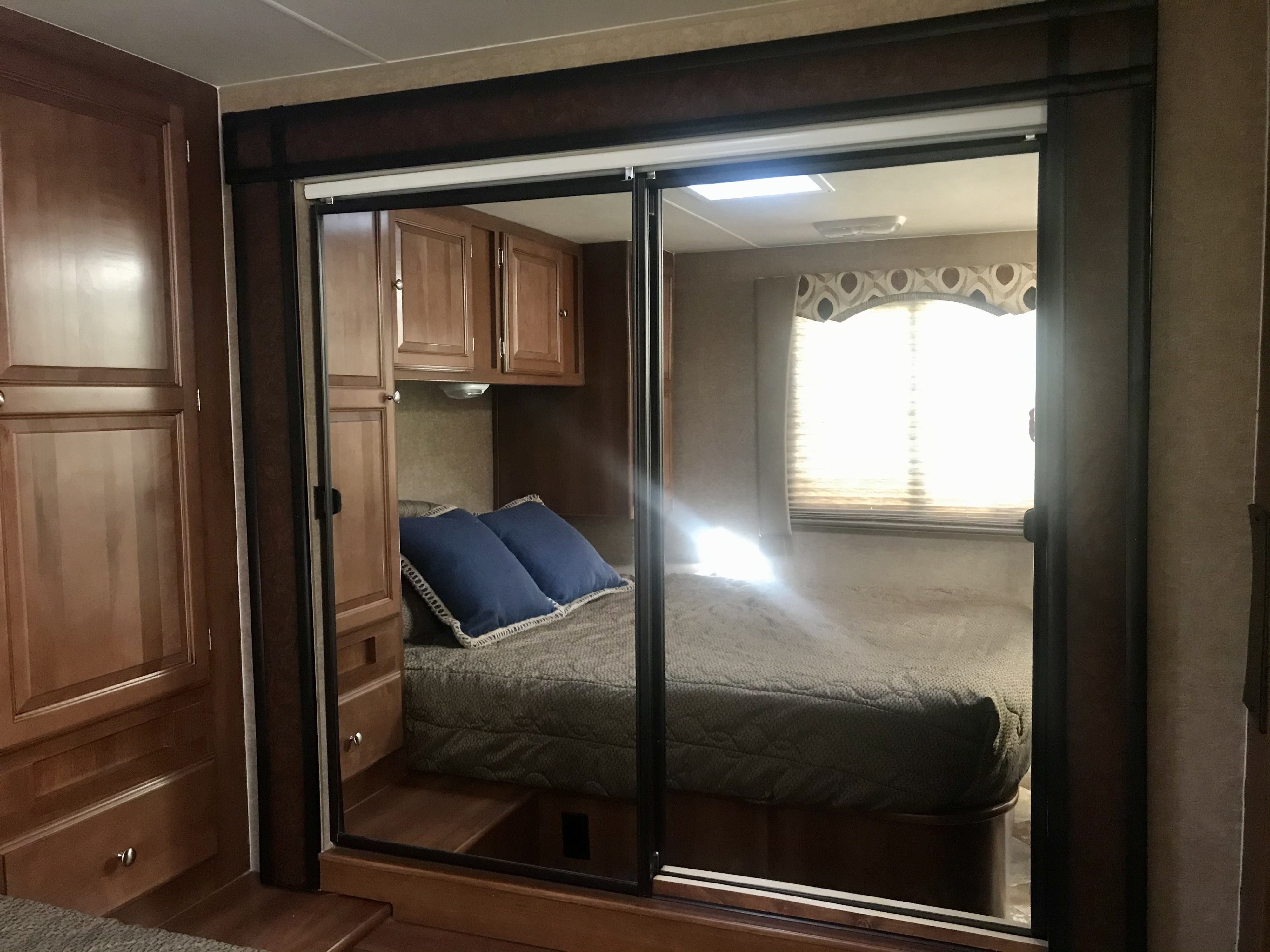 Inside this RV rental, a bedroom is seen through mirrored sliding doors, featuring a bed, bright window, and wood cabinetry.. Forest River Coachmen Leprechaun 2014