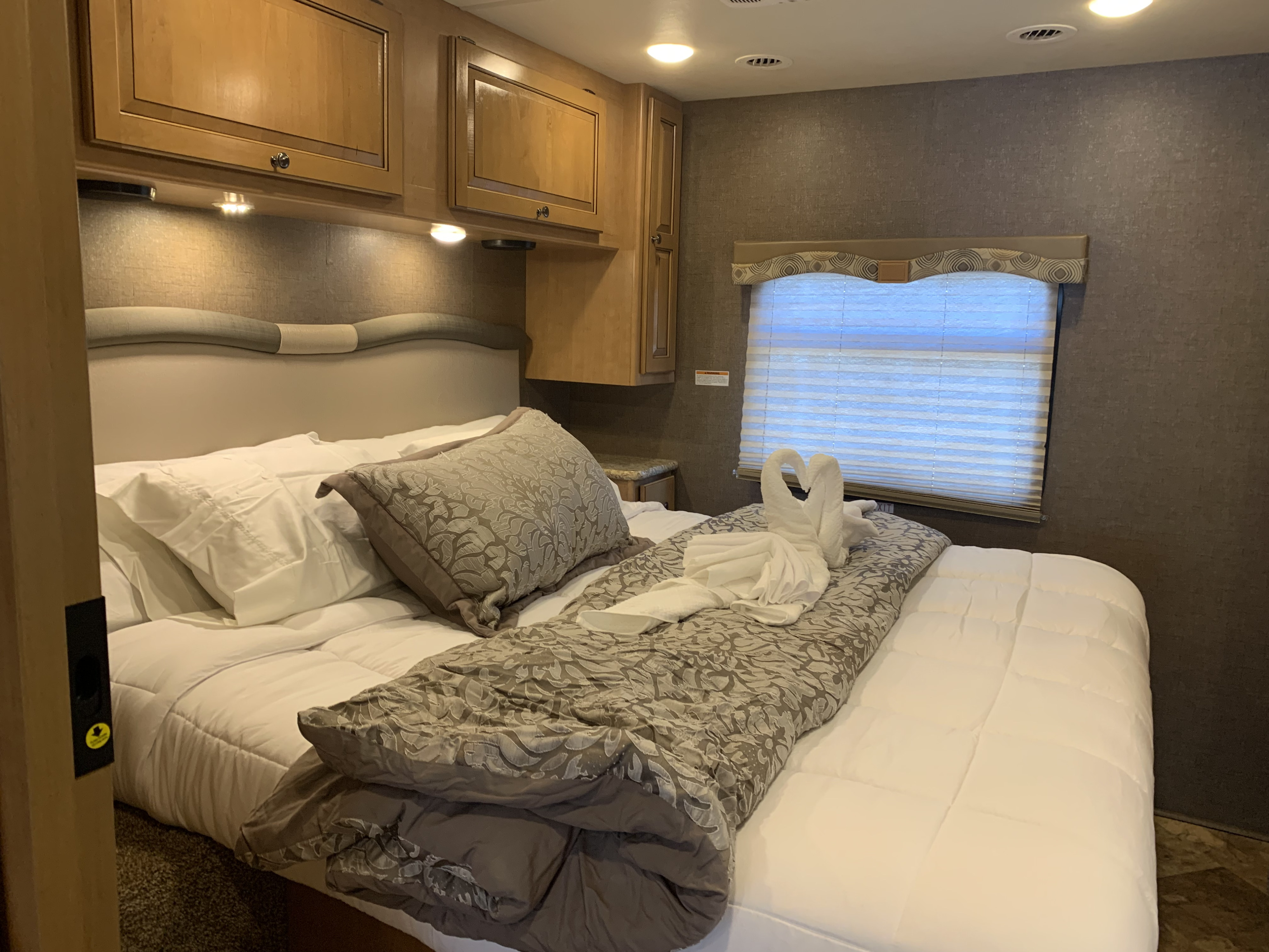 Inside an RV rental bedroom: a comfortable bed with swan towels, overhead cabinets, and a window with blinds.. Thor Windsport 2017