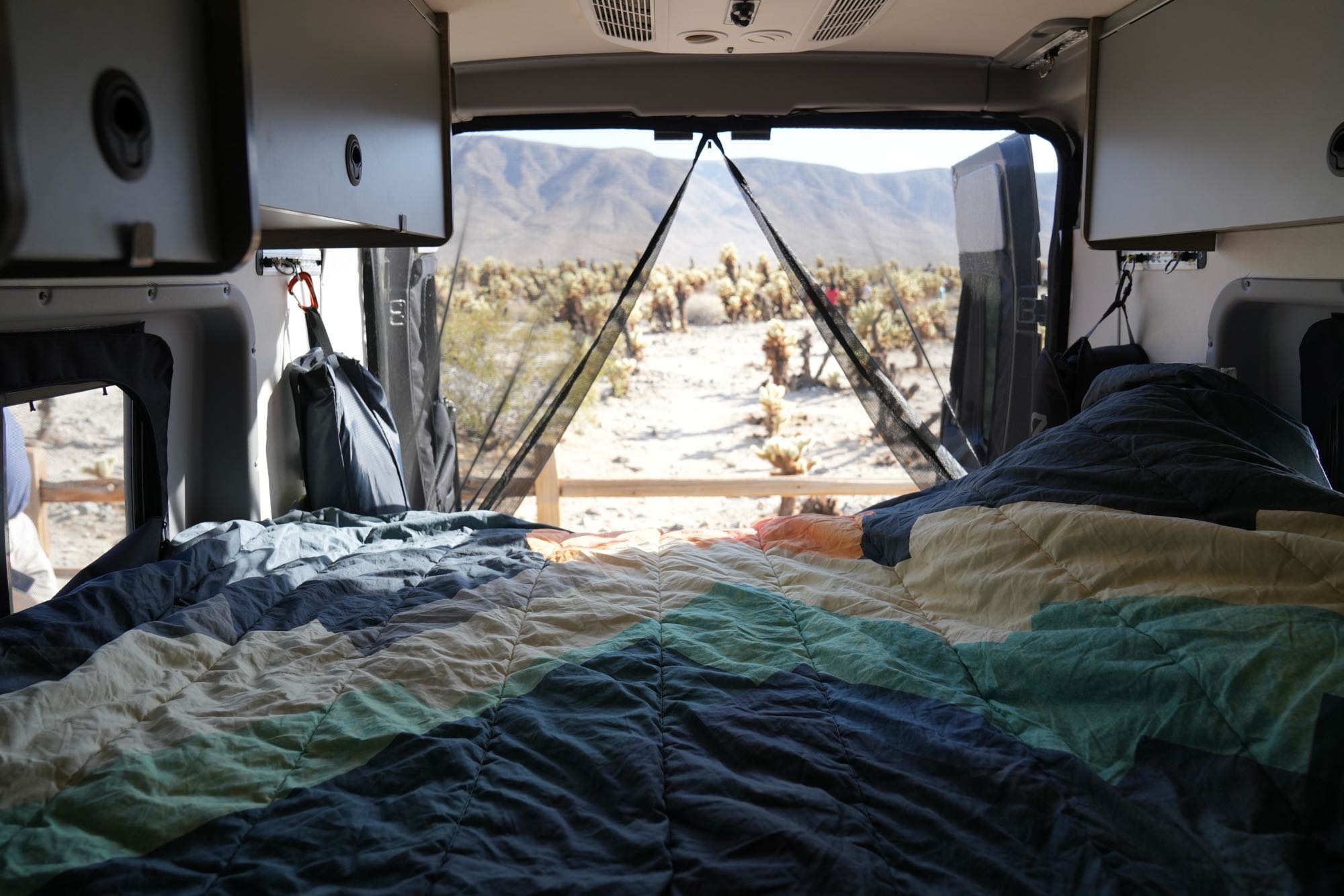 RV rental with a comfortable, patterned bed facing the open back, revealing a desert view of Cholla cacti and distant mountains.. Winnebago Solis 2022
