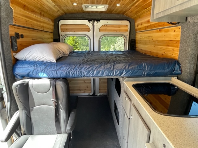 Cozy rv rental camper van interior with wood paneling. Raised bed, kitchen counter, and front passenger seat view.. RAM Promaster ALPINE X Edition OA15 2021