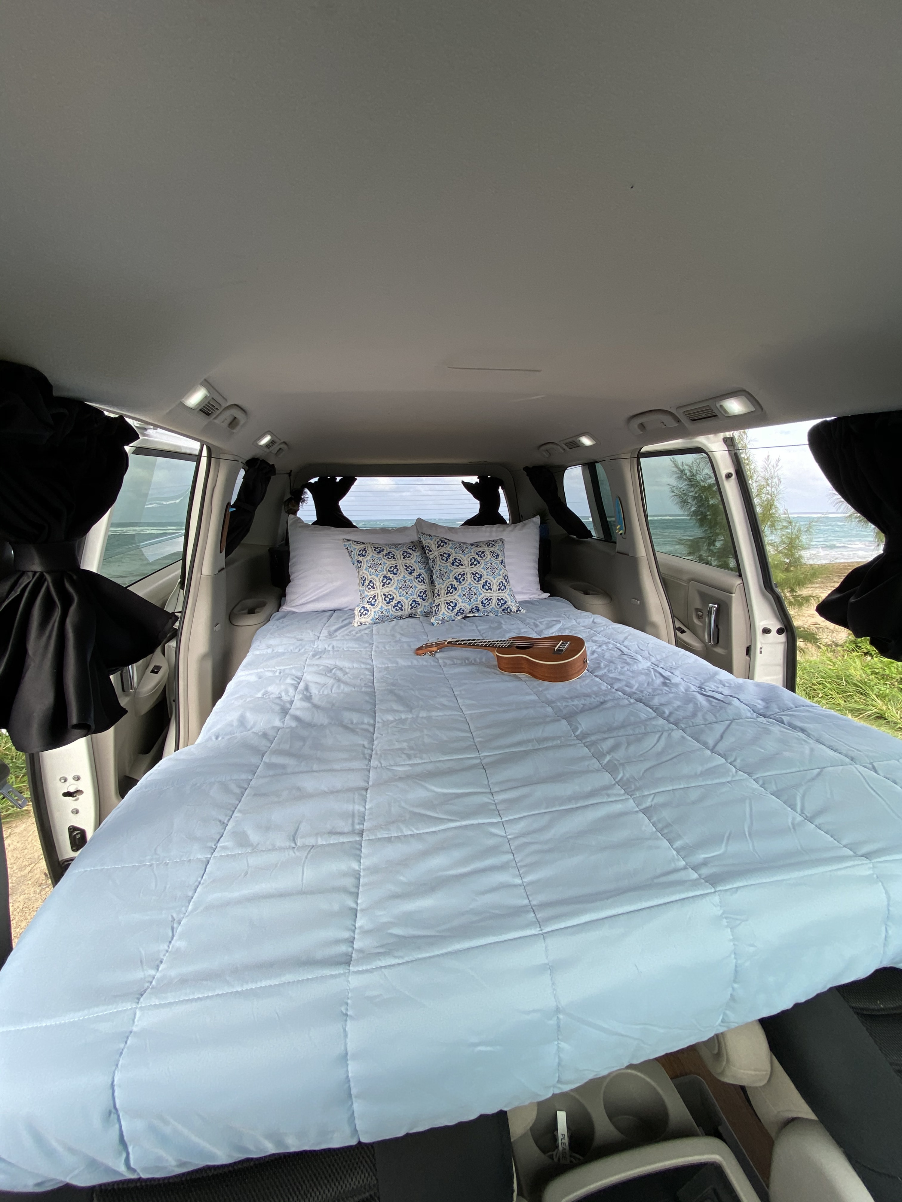 Inside an RV rental van: a comfy bed with a ukulele, offering panoramic ocean views. Ideal for a beachside adventure.. Nissan Quest 2017
