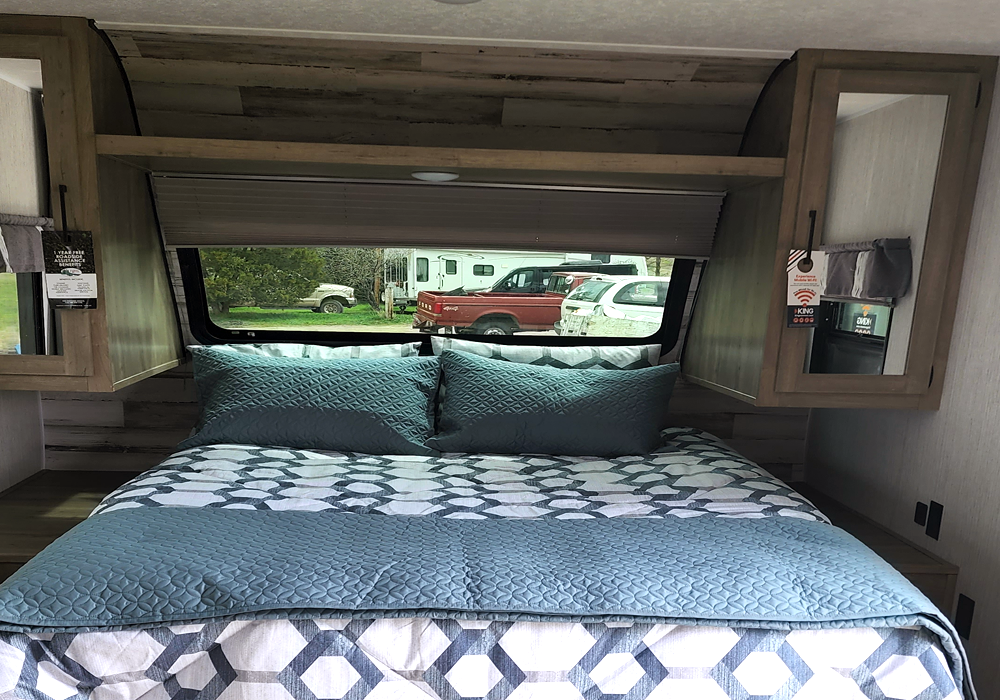 Cozy RV rental bedroom with blue bedding, overhead cabinets, and a window showing parked vehicles outside.. Forest River Puma 2021
