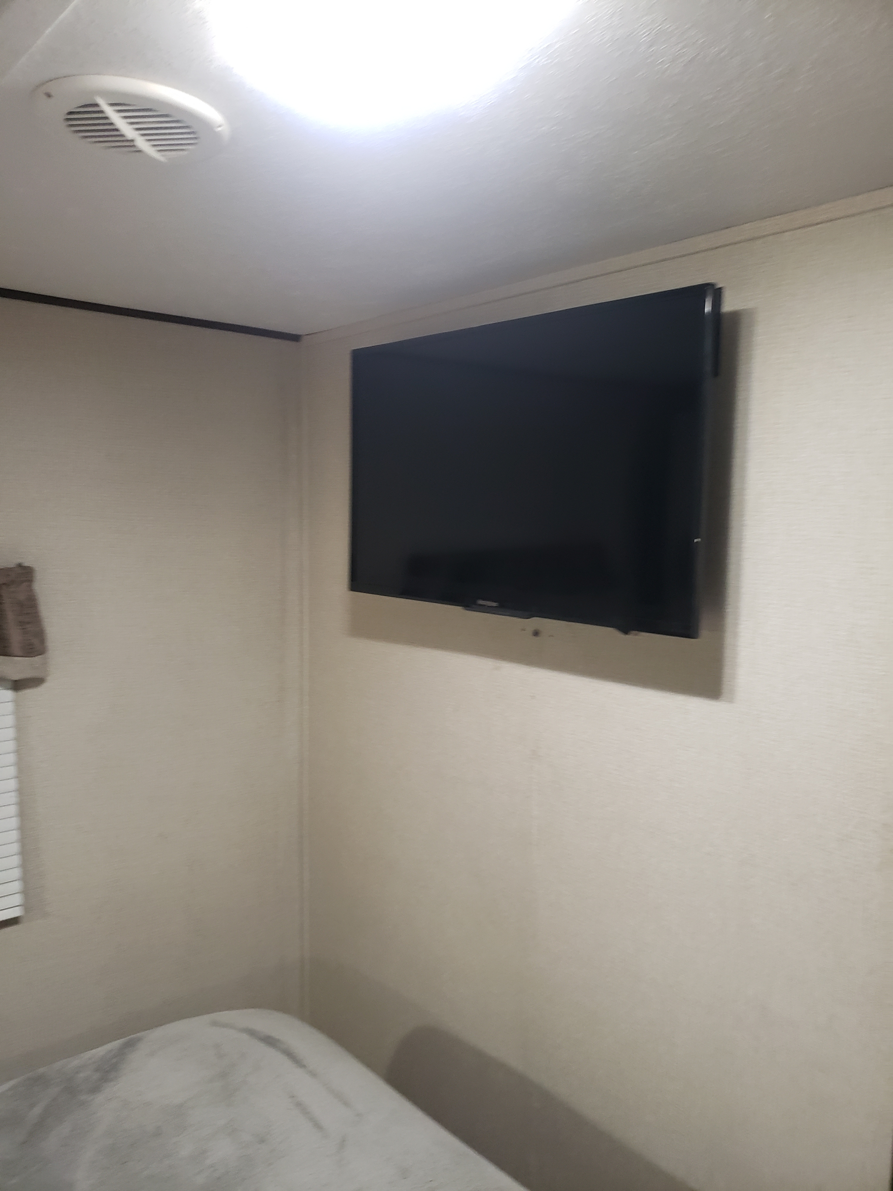 This rv rental interior showcases a wall-mounted TV, an adjacent bed, and a ceiling vent. Bright lighting illuminates the space.. Avenger ATI 2020