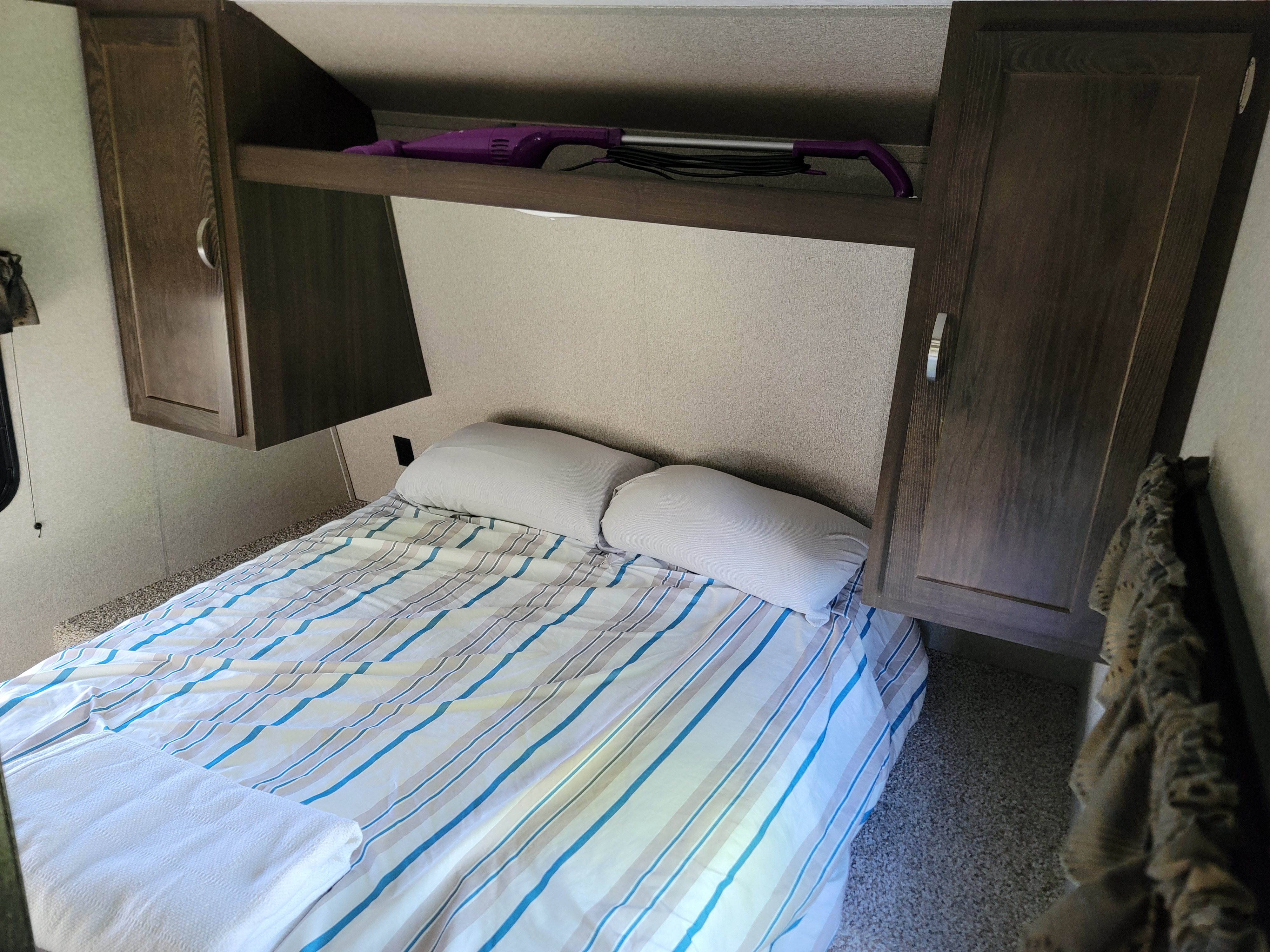Compact RV rental bedroom with striped bed, pillows, overhead storage, & purple vacuum.. Keystone RV Summerland 2018