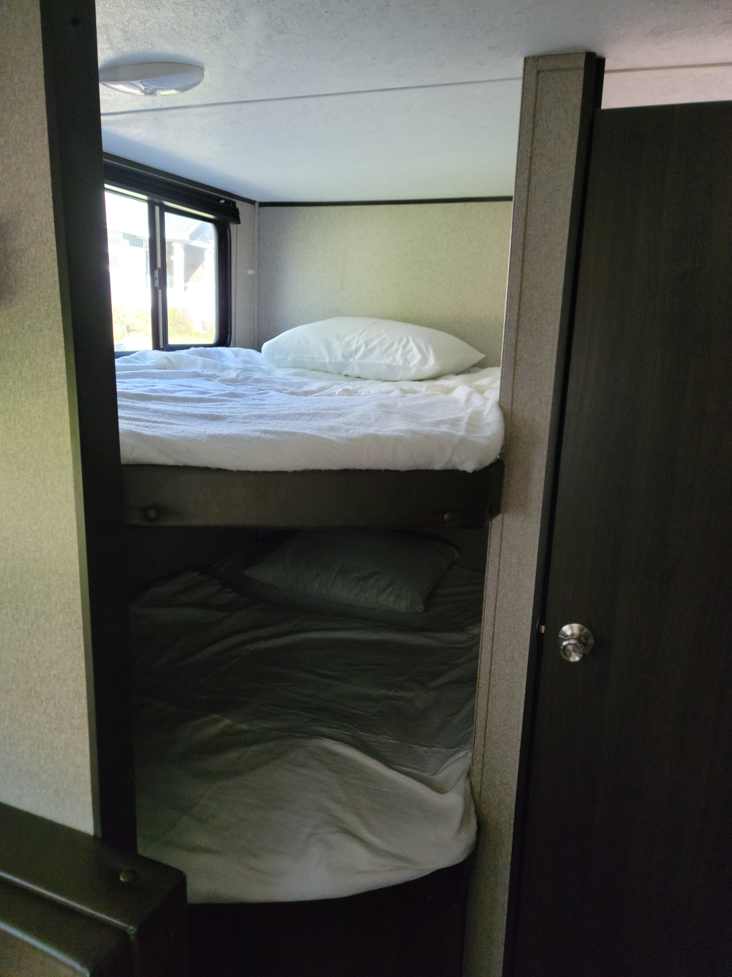 Inside an RV rental, a sleeping area features a bright upper bunk with white bedding and a cozy lower bunk with gray bedding, plus a window and door.. Keystone RV Summerland 2018