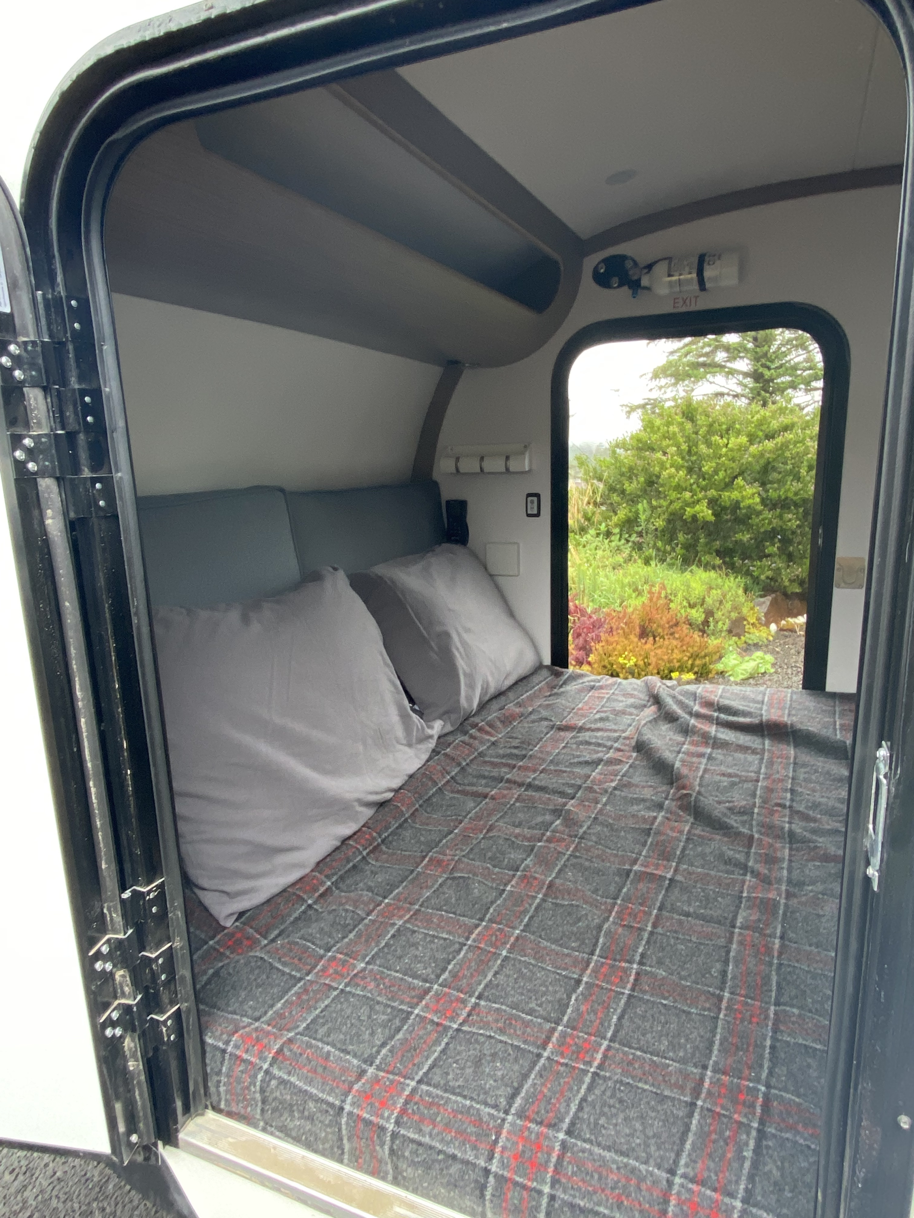 View into a compact RV rental: a bed with a gray plaid blanket, pillows, and a window showing lush outdoor greenery.. Bean Teardrop Trailer 2022