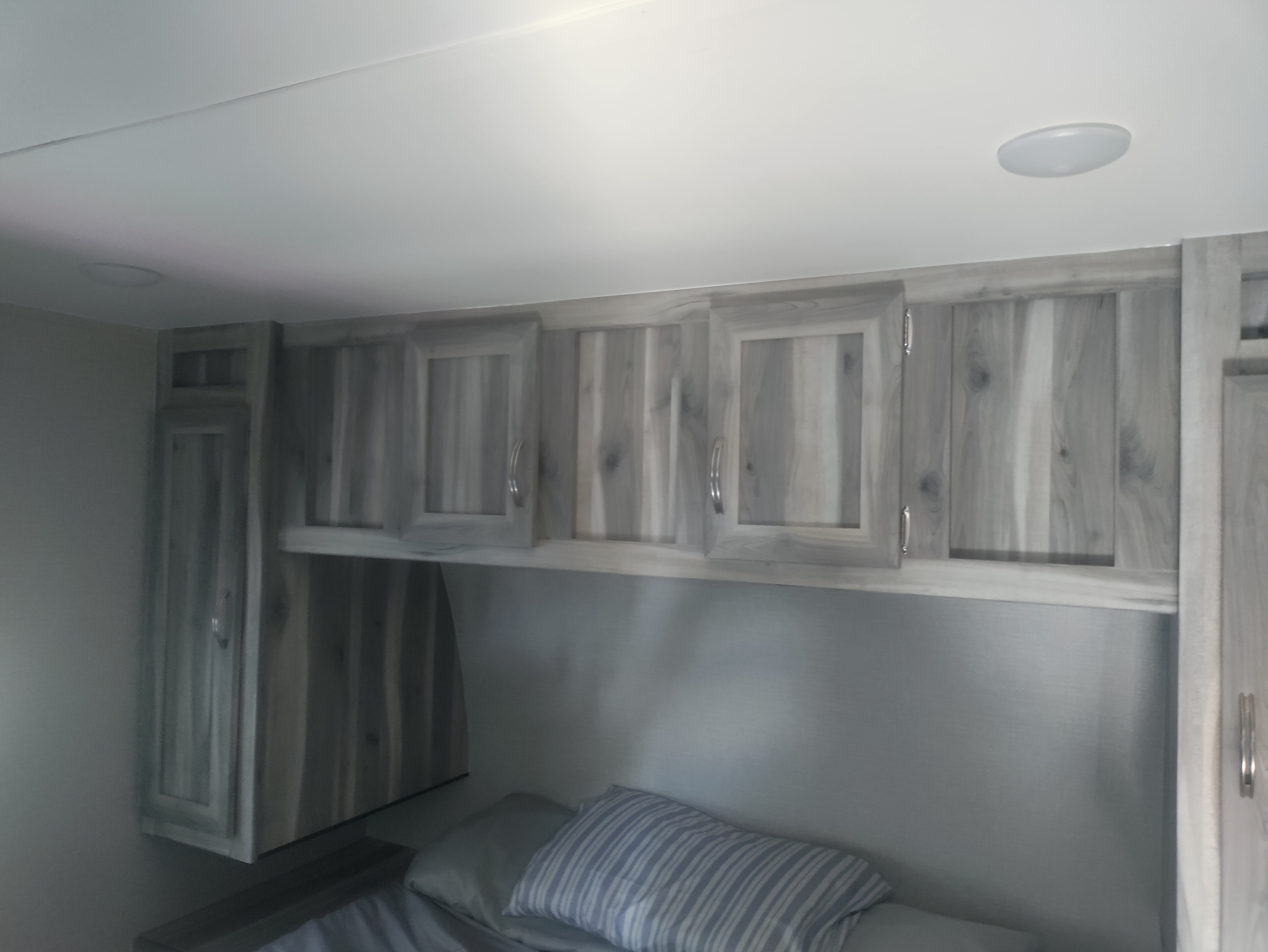 Cozy RV rental bedroom with light wood-grain overhead cabinets, a comfortable bed, and an integrated ceiling light.. Chinook Dream bunkhouse 2022
