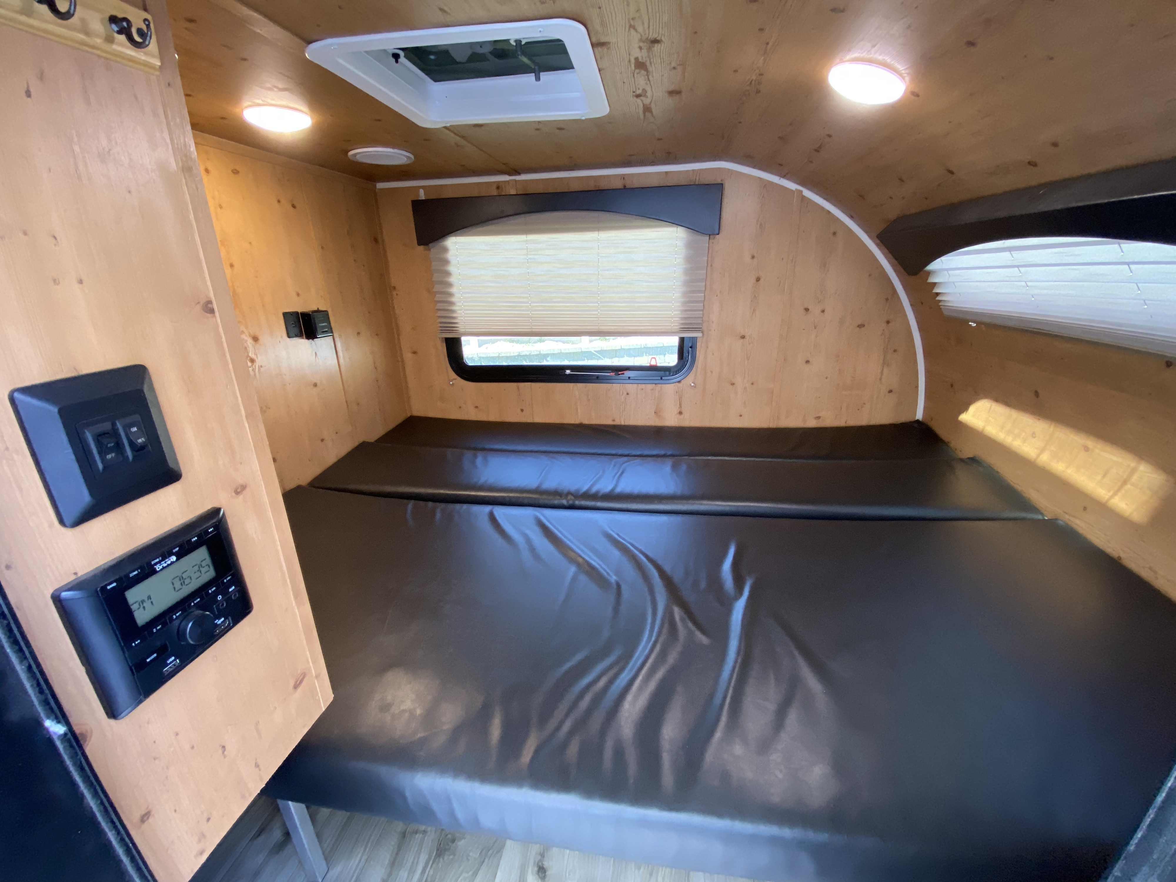 Cozy RV rental interior shows a black padded bed, wooden walls, a window with blinds, and built-in controls.. Sunset Park RV SunRay 2020