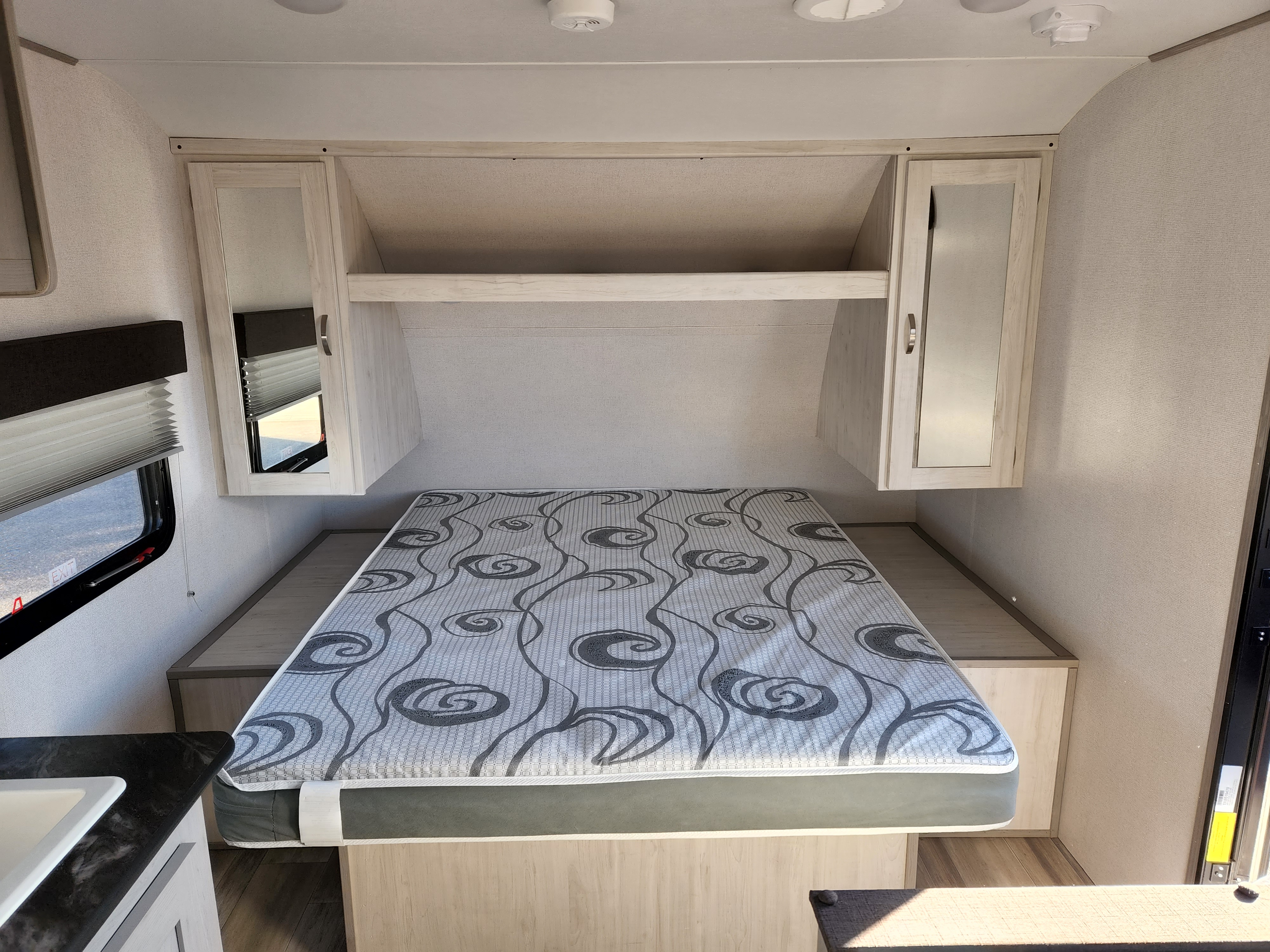 Inside an RV rental: a compact bedroom with a patterned mattress, light wood overhead cabinets, and a window with blinds.. Prime Time Tracer 2021