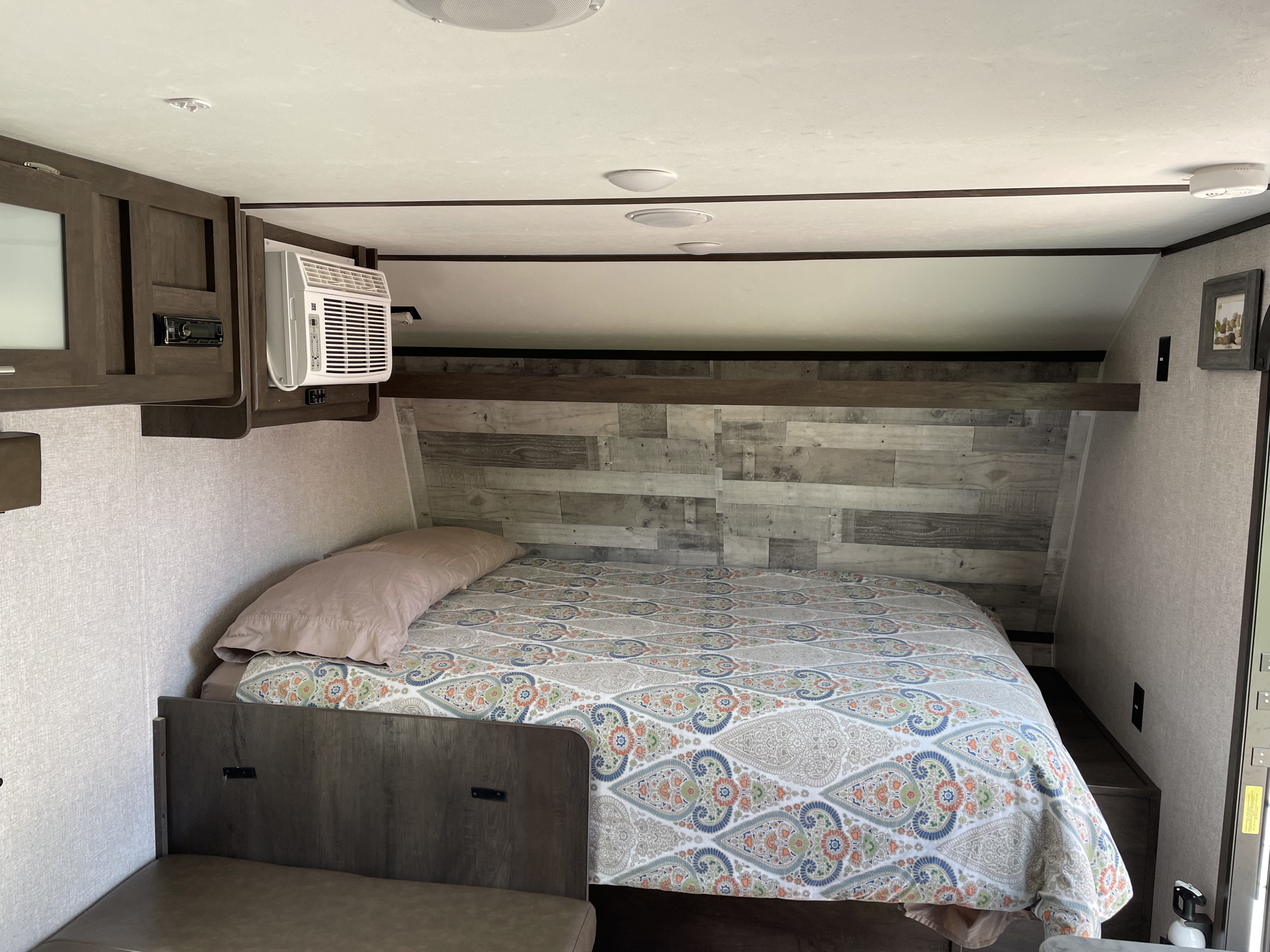 A cozy RV rental bedroom featuring a patterned bed, window AC, and rustic wood paneling for comfort on the road.. Dutchmen Coleman Lantern LT 2020