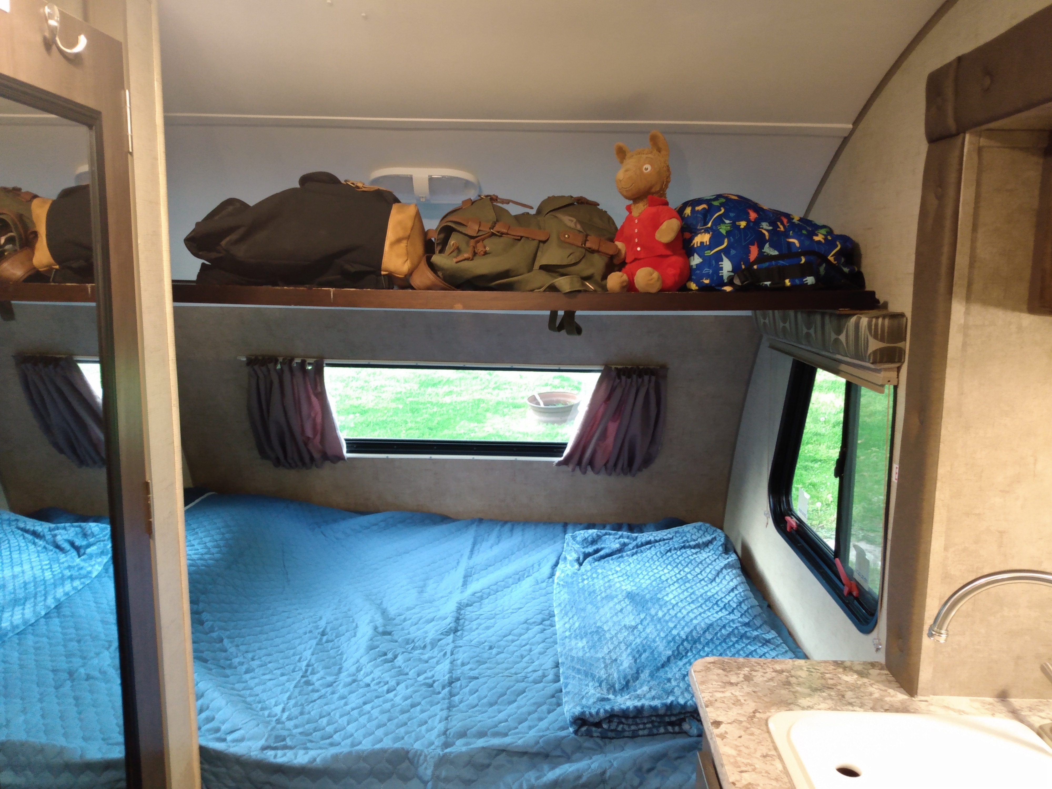 RV rental interior: bunk storage with bags, lower bed with blue quilt, outdoor window view, and sink.. R-Pod R-Pod Trailer 2015