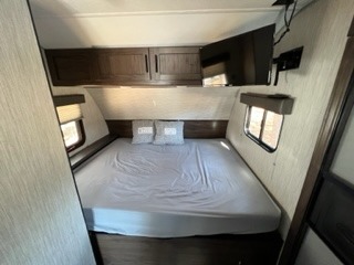 An RV rental's cozy bedroom, featuring a large bed with light sheets, overhead wood cabinets, and a mounted TV, ready for travel.. Forest River Cherokee Wolf Pup 2021