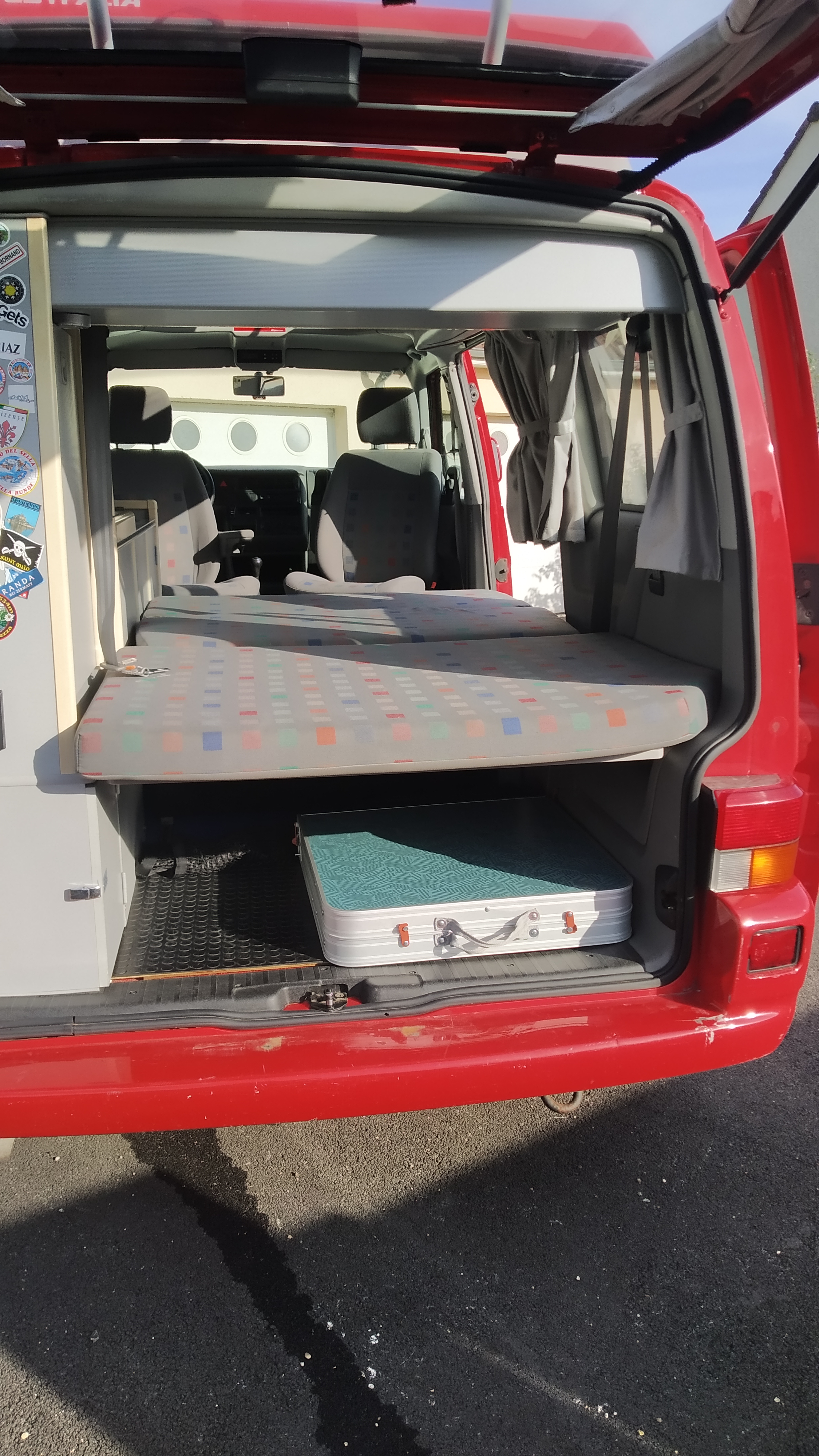 Rear interior of a red camper van RV rental, showing a patterned bed, lower storage with a folded table, and open hatch.. Volkswagen CALIFORNIA 2002