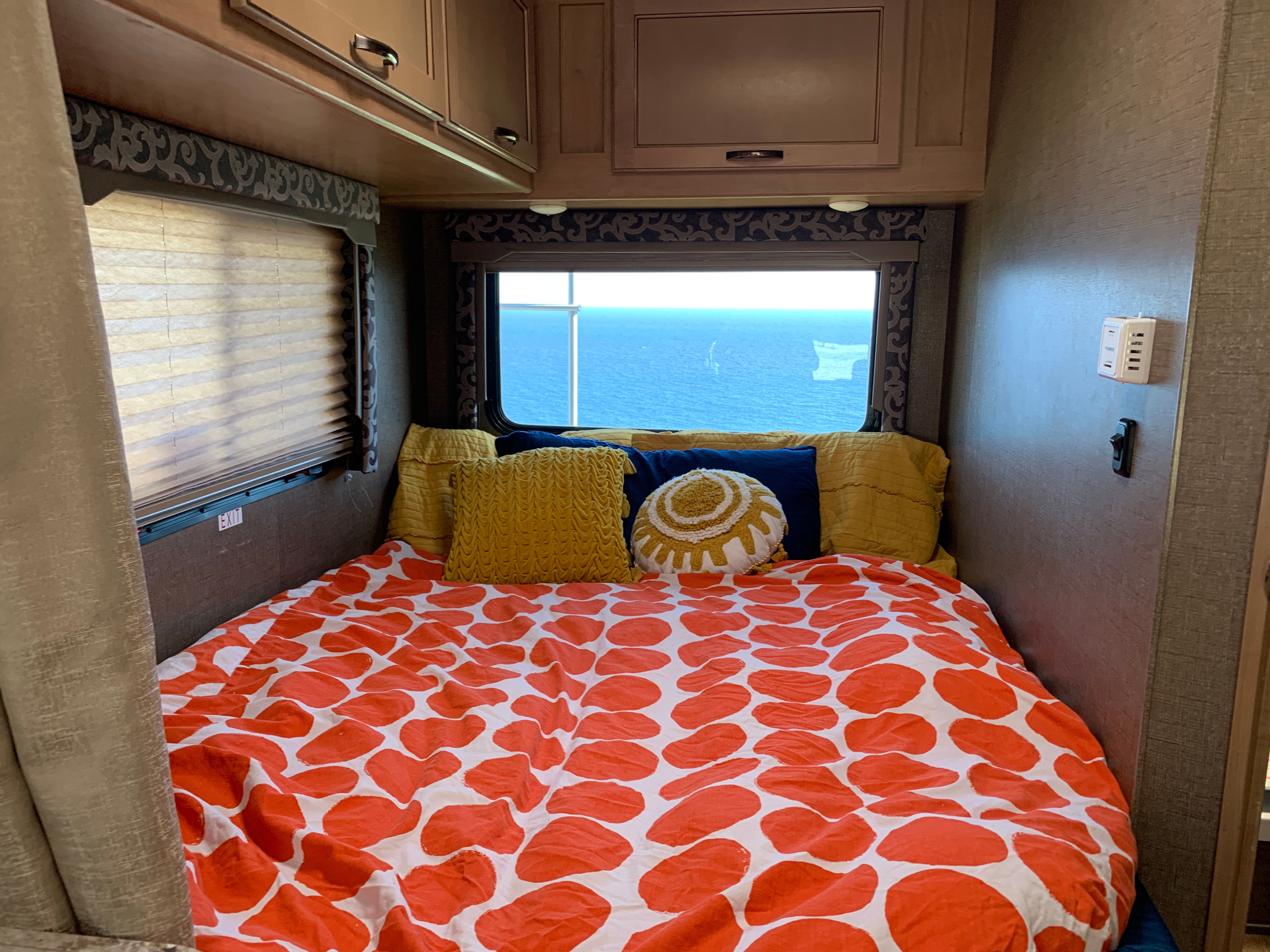 Cozy RV rental bedroom featuring a bed with orange duvet, yellow pillows, and a stunning ocean view from the window.. Thor Four Winds 2020
