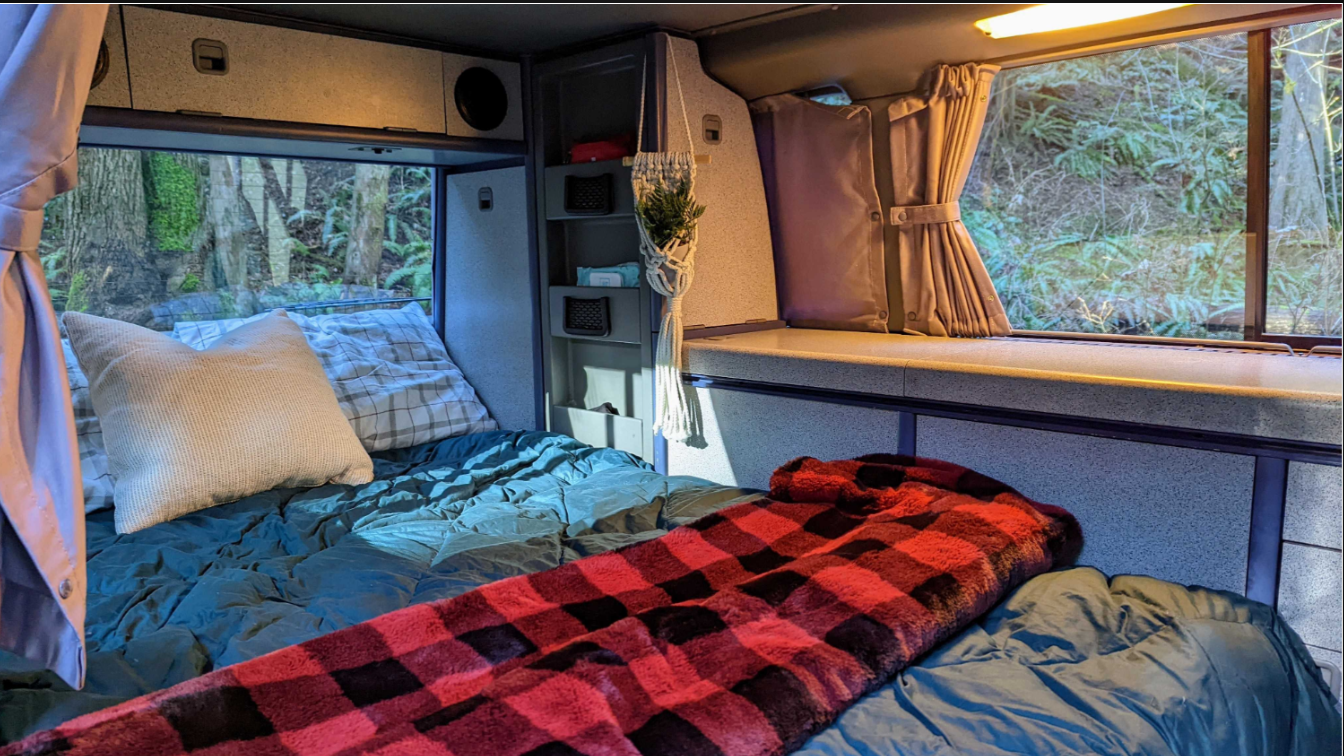 Inside a comfy RV rental: bed with plaid blanket, built-in storage, and lush forest views. Great for adventures.. Volkswagen T4 Westfalia 1993