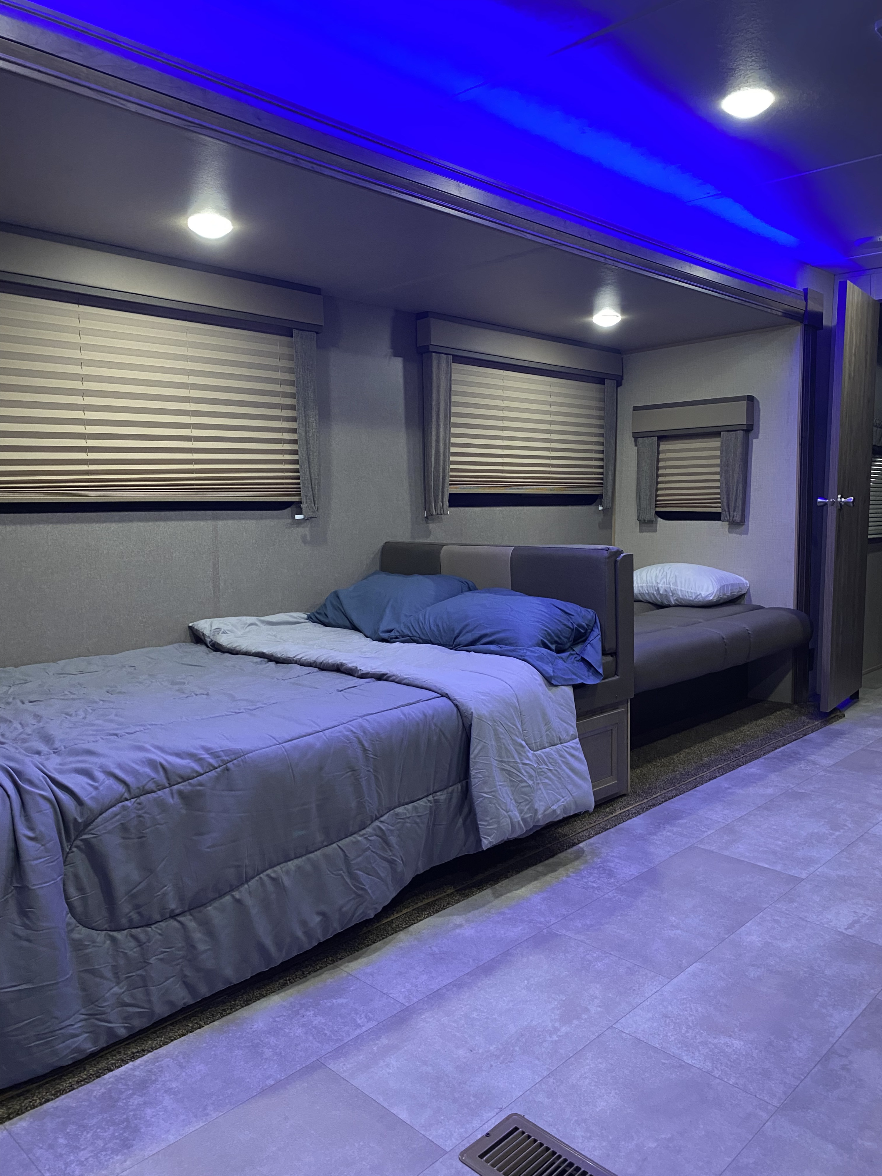 Inside an RV rental: a modern bedroom featuring two beds with blue linens, white and blue lighting, and window blinds.. Jayco Jay Flight 2021