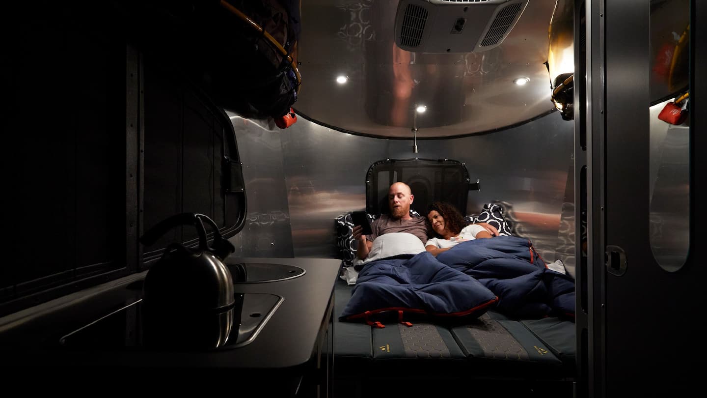 Cozy RV rental interior with a couple relaxing in bed; the man uses a tablet, kettle visible on the counter.. Airstream Base Camp 2023