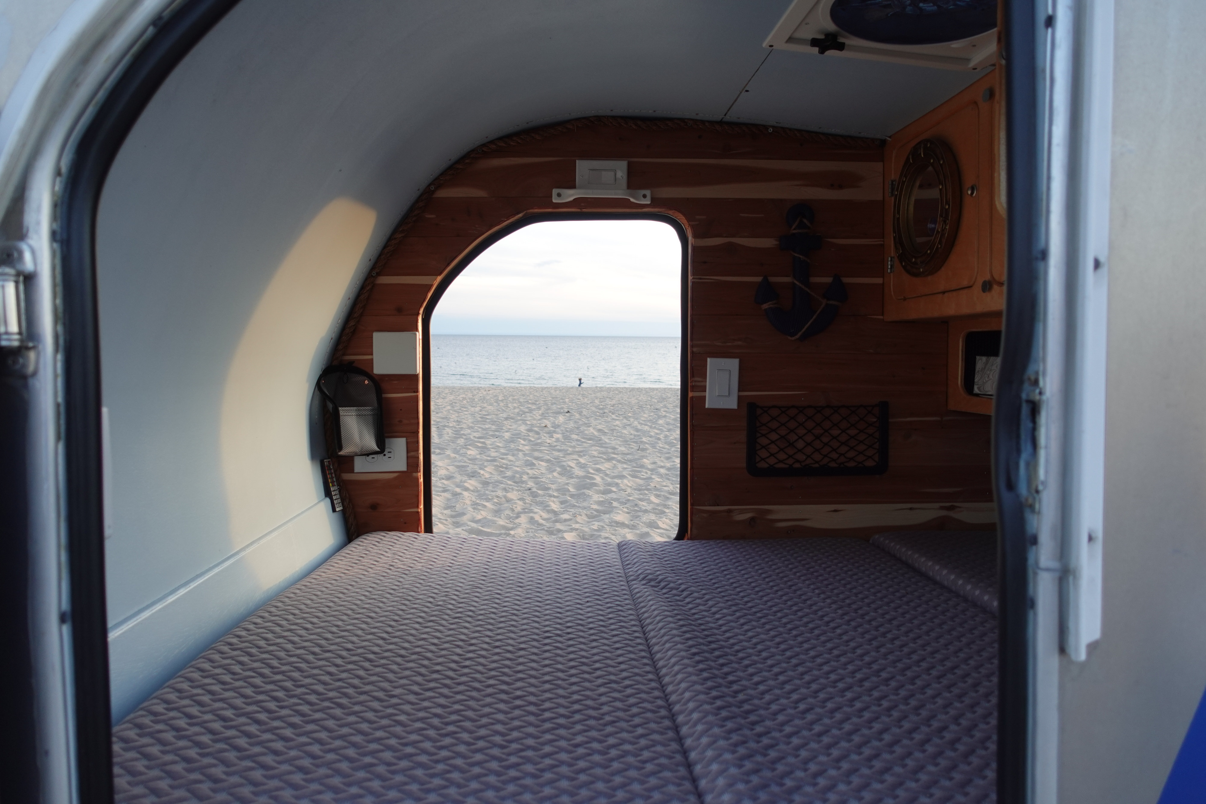 Cozy rv rental interior with bed and wood details, looking out to a serene sandy beach and calm ocean.. Second Wind Freedom 2020