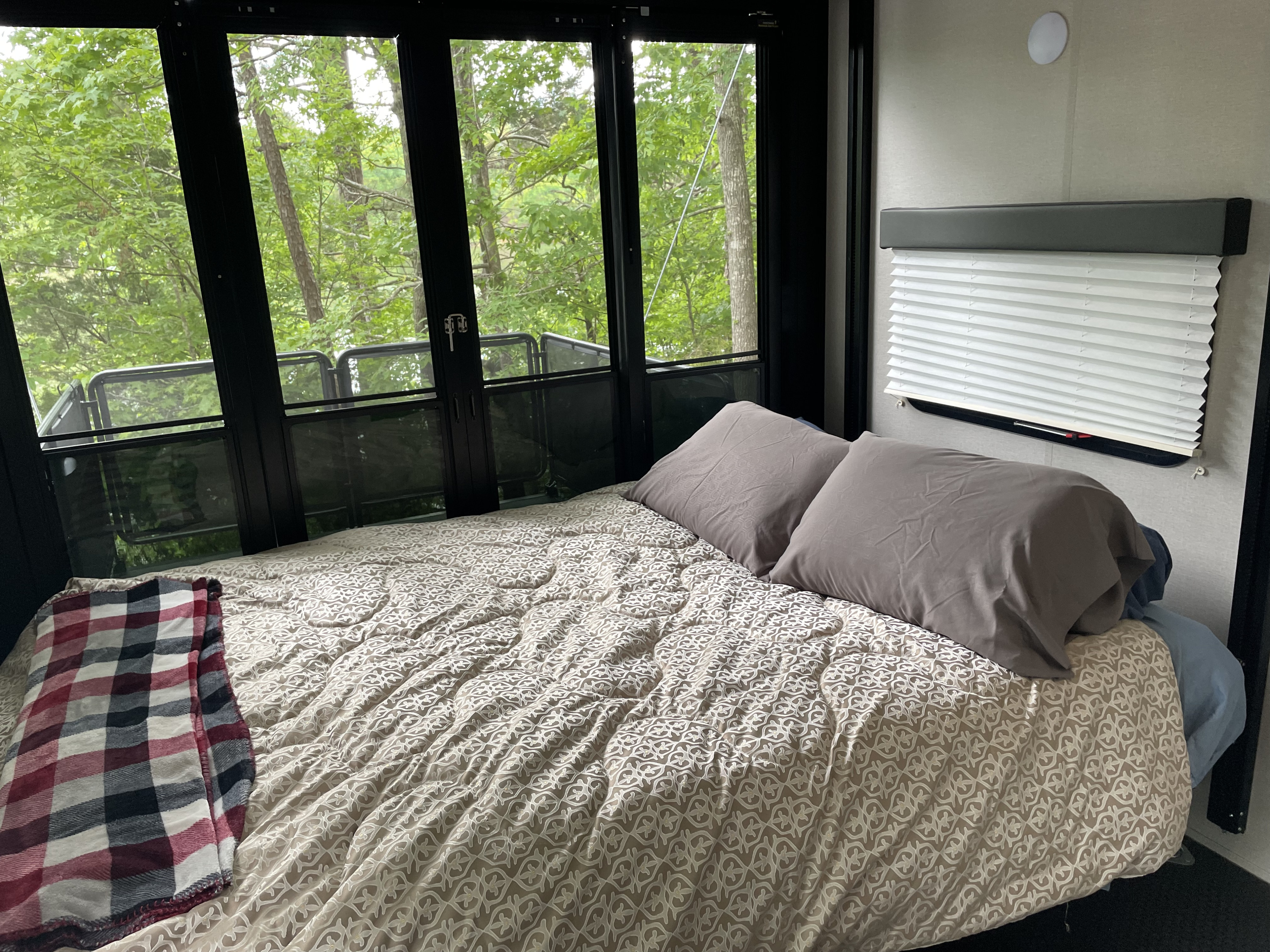 Cozy RV rental bedroom: a comfortable bed with gray pillows, plaid blanket, and large windows with a forest view.. Grand Design Momentum Toy Hauler 2022