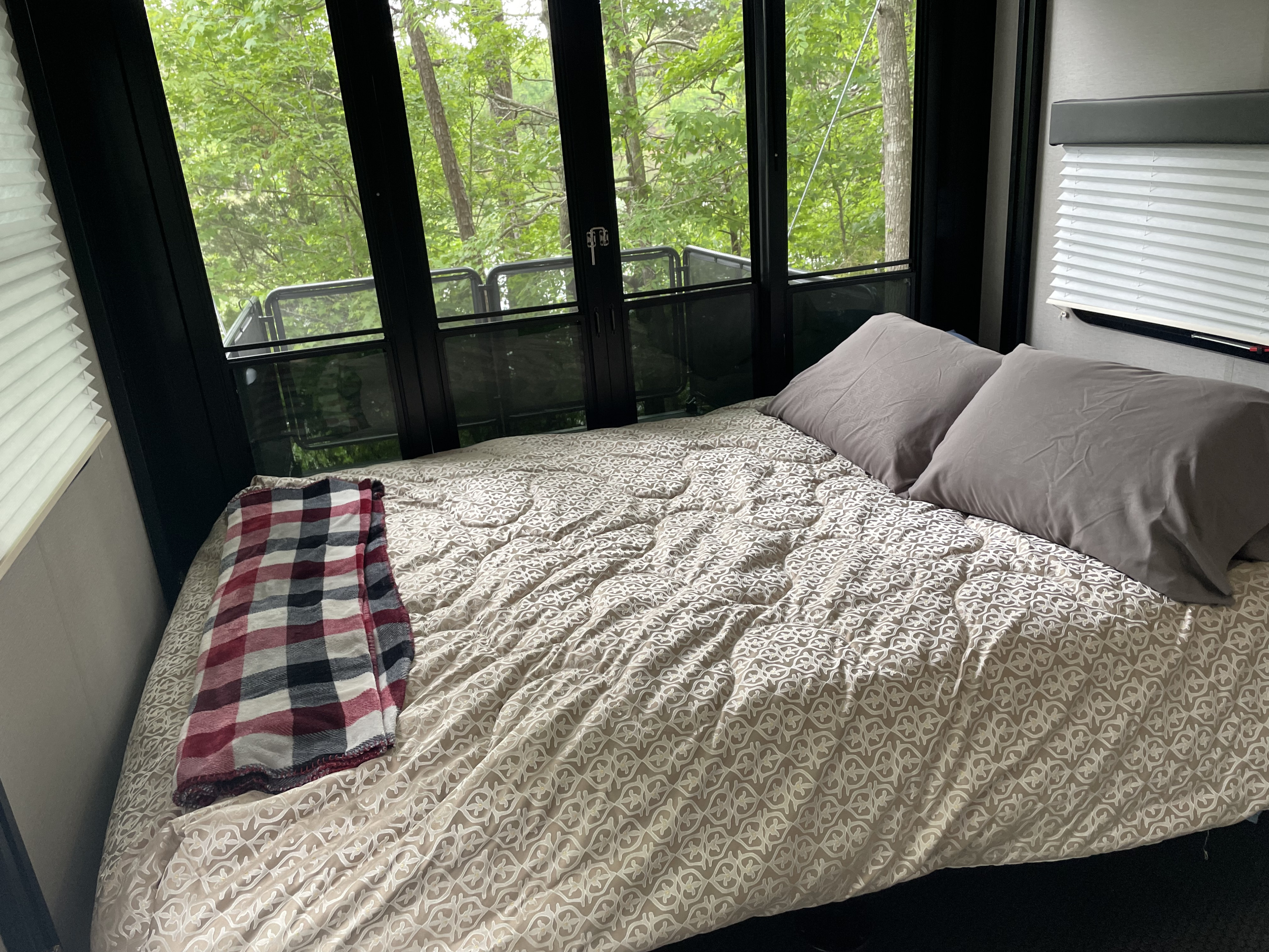 Serene RV rental bedroom with a comfy bed, plaid blanket, and expansive windows offering a beautiful forest view.. Grand Design Momentum Toy Hauler 2022