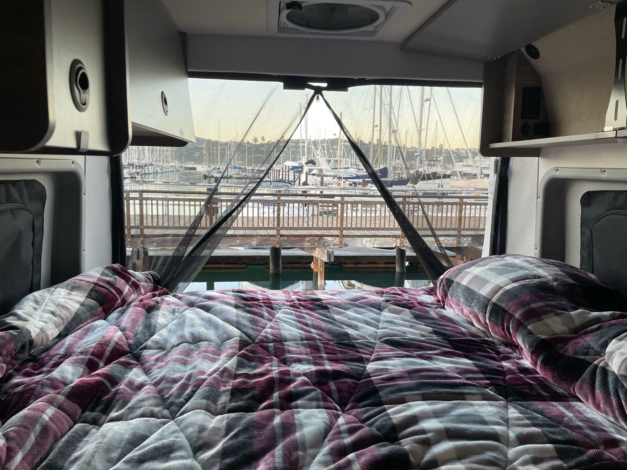 Relaxing in an RV rental, looking out at a stunning marina. Cozy plaid bed, boats, and docks create a serene waterfront view.. Winnebago Winnebago Solis 59P 2022