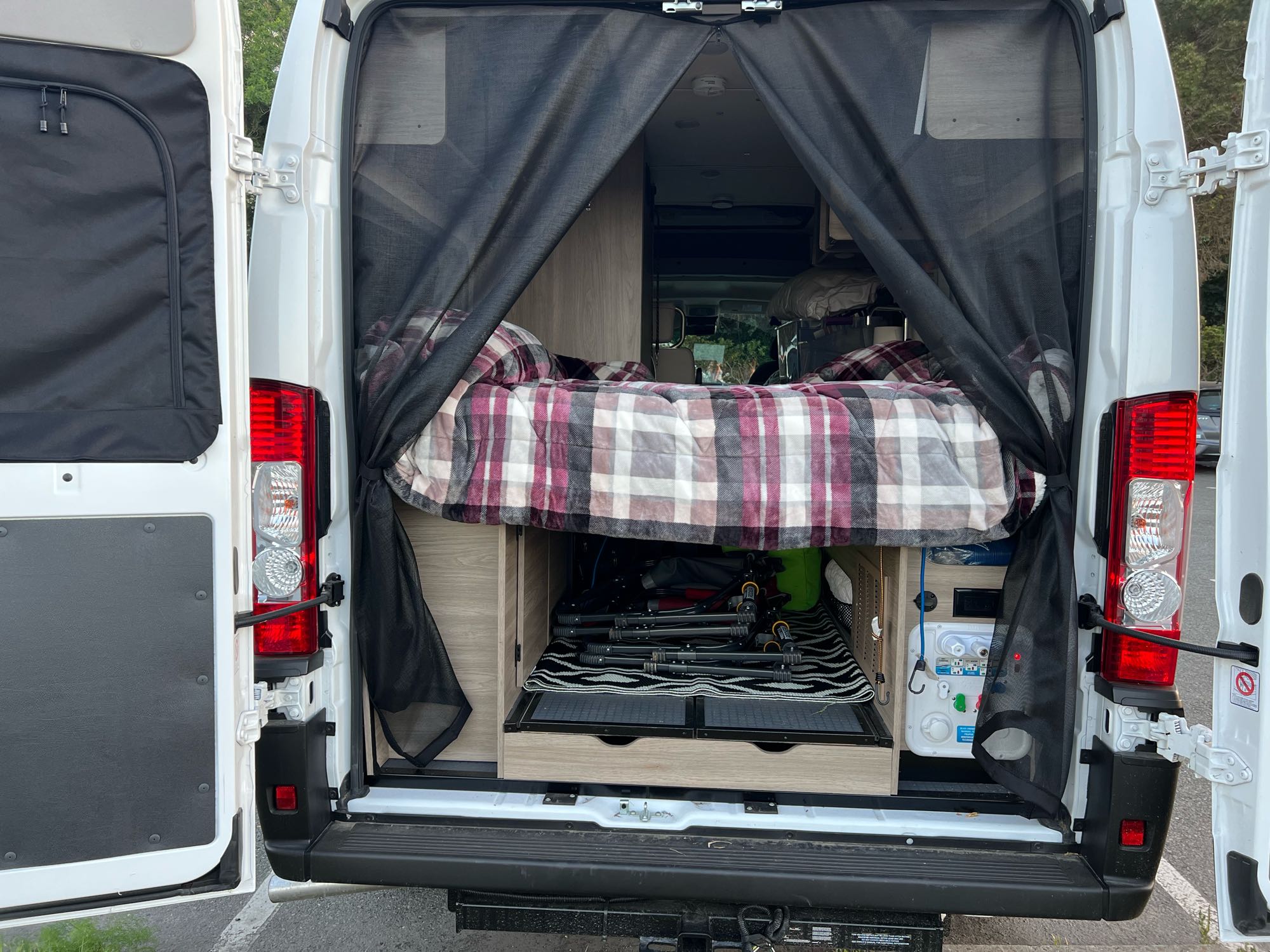 RV rental interior: cozy plaid bed, organized under-bed storage, & bug screen seen through open rear doors. Mobile home.. Winnebago Winnebago Solis 59P 2022